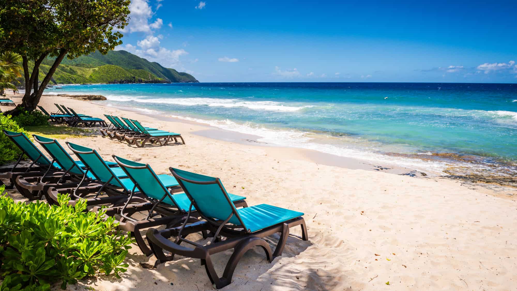 A row of turquoise lounge chairs sits on a white sand beach, facing the crystal-clear ocean waves. Lush green hills rise in the background under a bright blue sky.