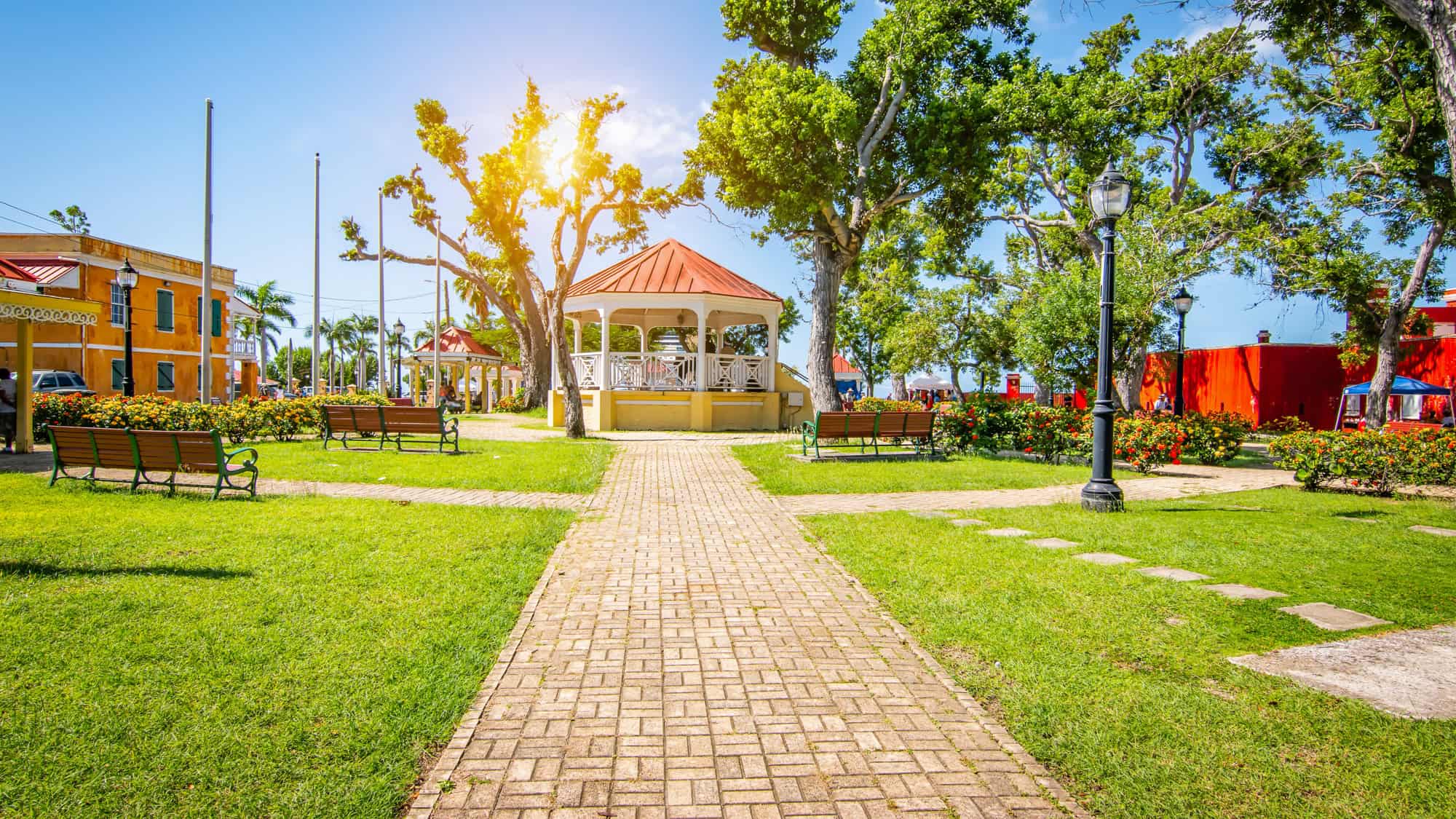 A scenic park with a well-manicured lawn, bright flowers, and a charming white gazebo with a red roof. Sunlight filters through tall trees, casting shadows on the pathways.