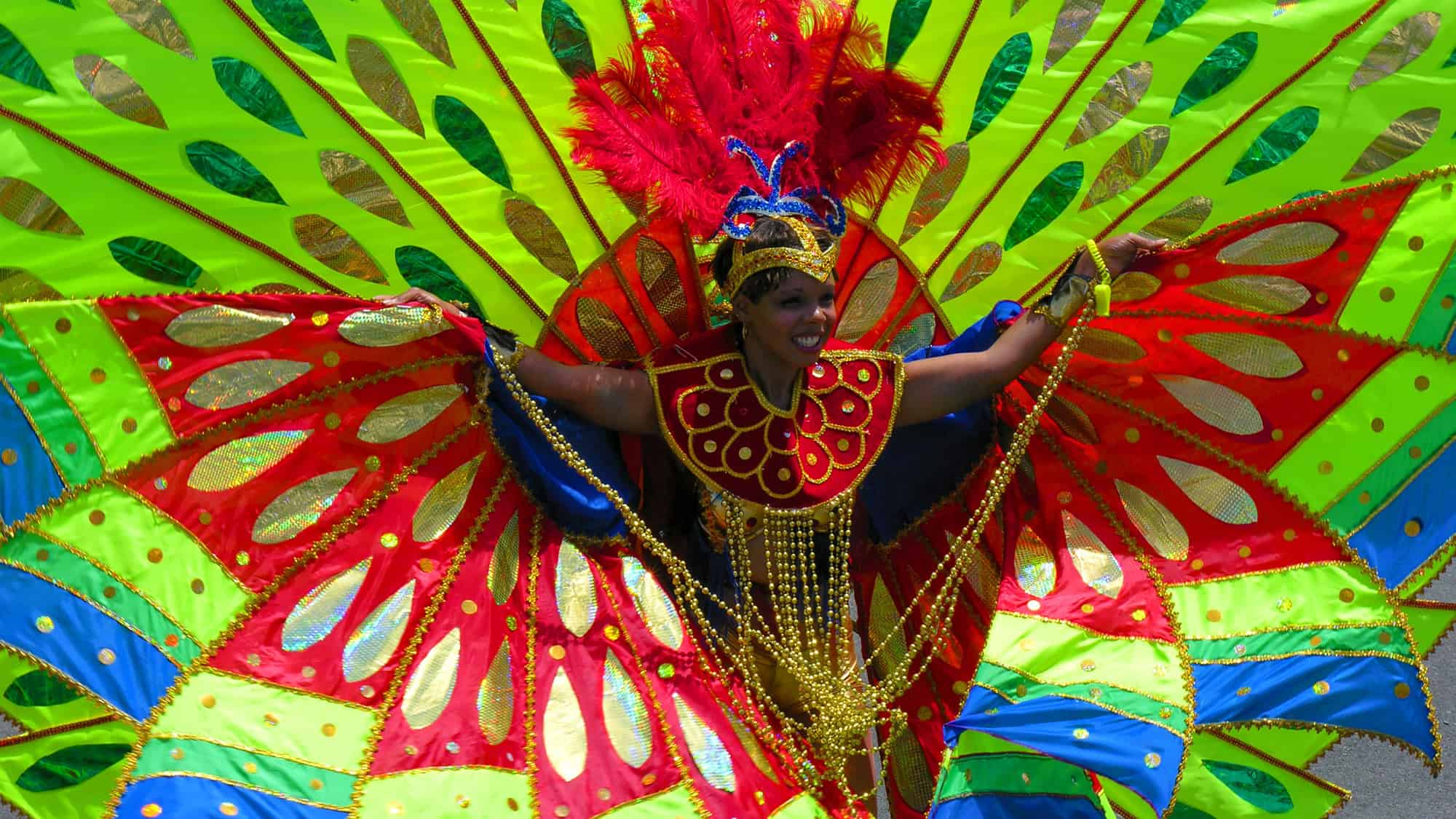 A vibrant performer in an elaborate costume spreads a massive, fan-like display of neon green, red, and gold fabric. She smiles joyfully, surrounded by festival energy.