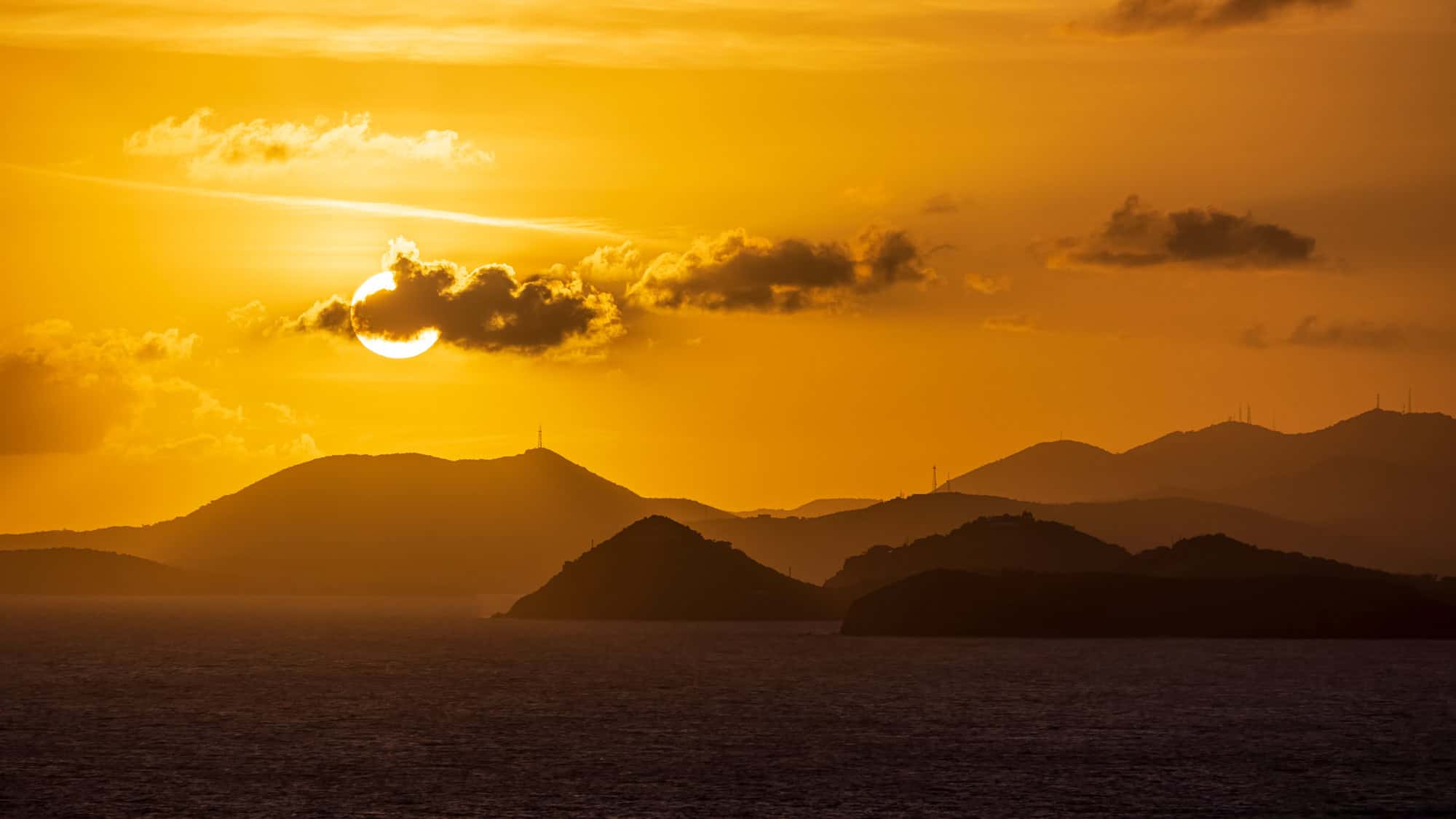 A golden sunset casts a warm glow over the Virgin Islands, with the sun partially hidden by a cloud. The silhouetted mountains and islands in the distance contrast beautifully against the fiery sky.