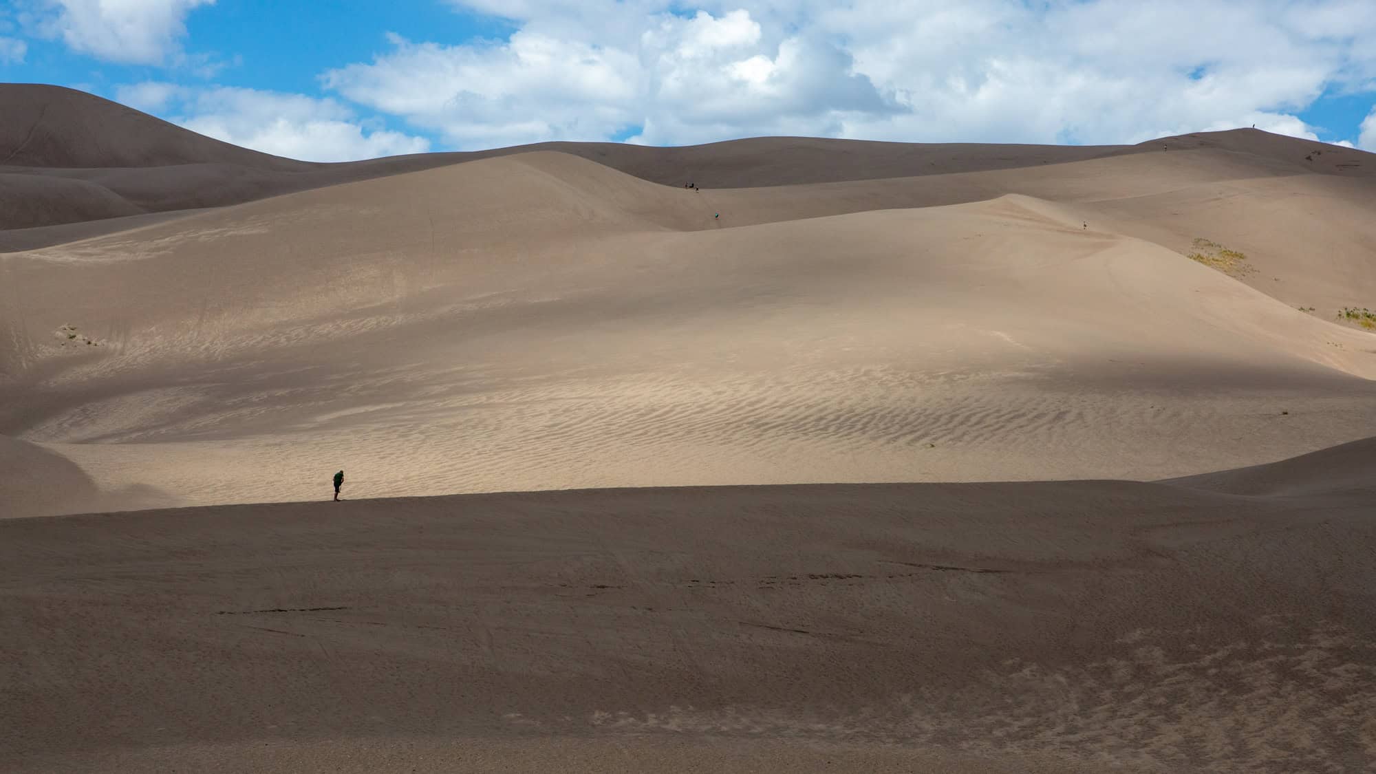 A lone hiker crosses a massive sand dune under a partly cloudy sky, dwarfed by the sweeping, textured hills of the Great Sand Dunes in Colorado.