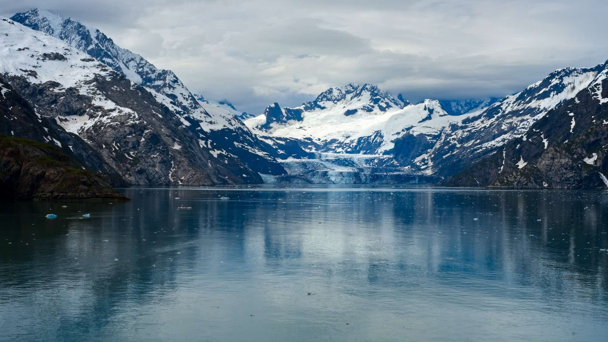 A dramatic Alaskan landscape featuring towering snow-capped mountains and a calm bay dotted with floating ice, reflecting the rugged peaks above.