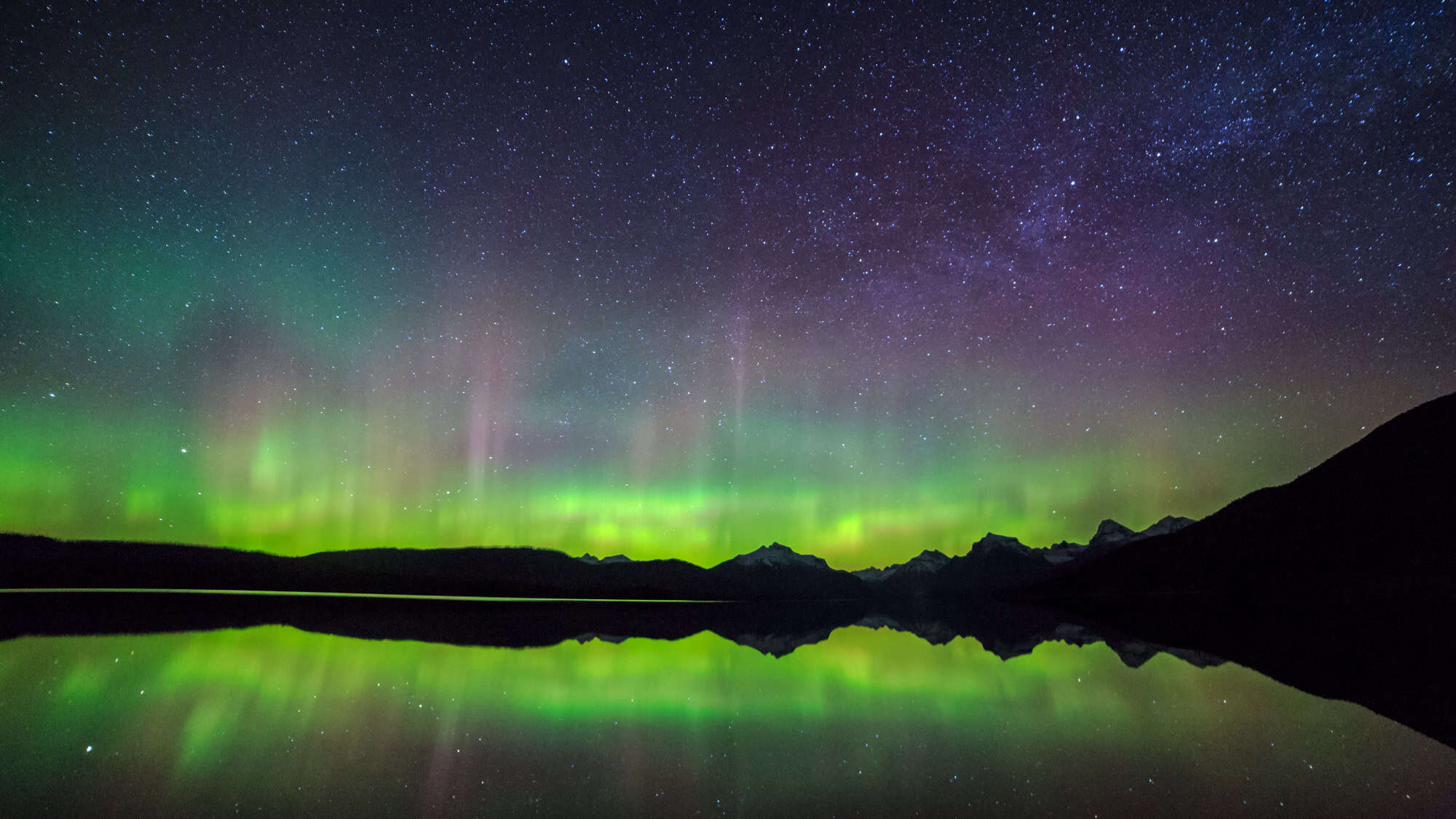 Green and pink aurora streaks shine over snowy mountains and trees, reflecting in a small pond beside a snow-covered trail.