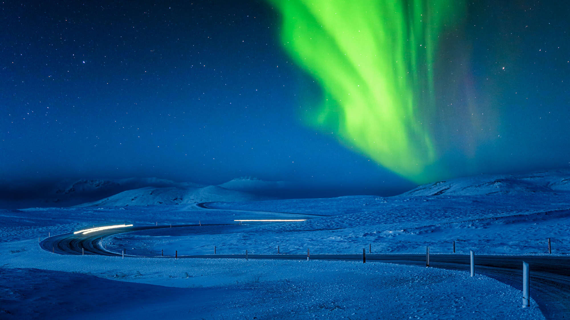 A vivid green aurora swirls across a deep blue sky above a winding snow-covered road illuminated by car lights.