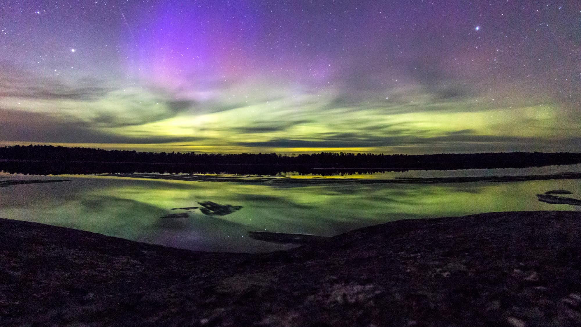 Waves of green and purple aurora ripple through the sky and reflect across a still lake at night.