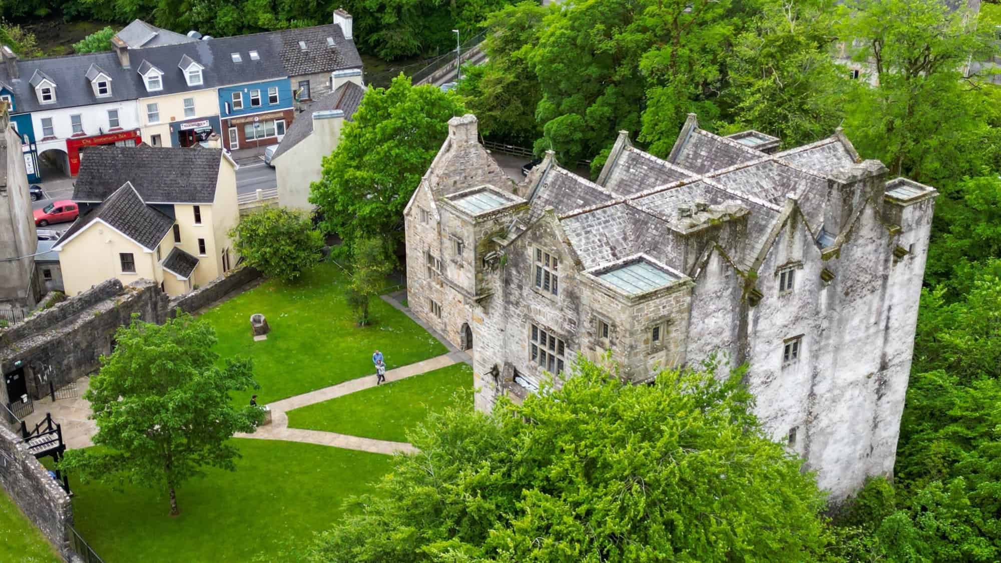 Aerial view of Donegal Castle in Ireland, showcasing its gray stone walls, gabled rooflines, and glass skylights surrounded by lush green trees. The foreground includes a manicured lawn with walking paths and a few visitors near the entrance.