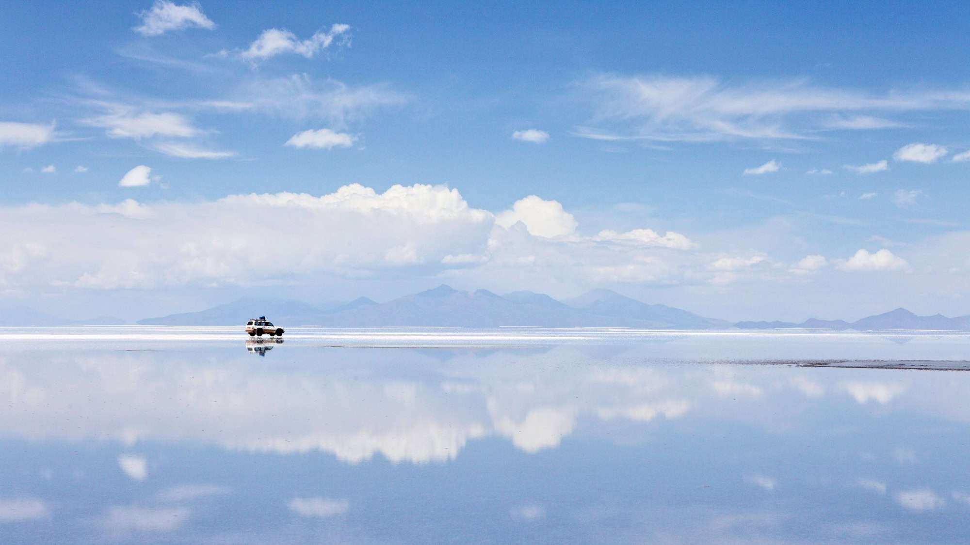 A lone SUV reflected on the expansive, mirror-like surface of the Salar de Uyuni salt flats in Bolivia, under a vast blue sky with distant mountains.