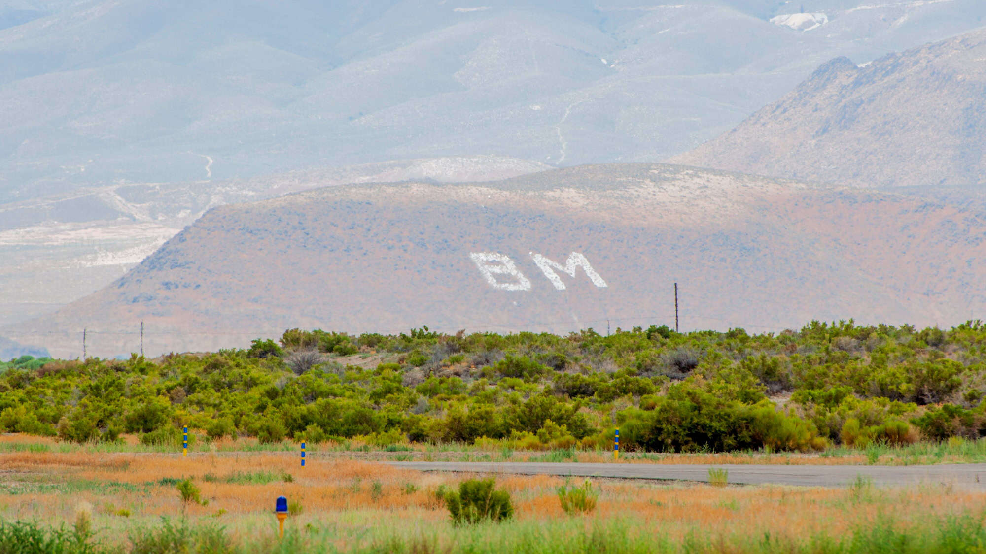 A hillside near Battle Mountain, Nevada, features large white-painted "BM" initials, commonly seen on western U.S. hillsides as town markers, with a sagebrush-covered plain in the foreground.