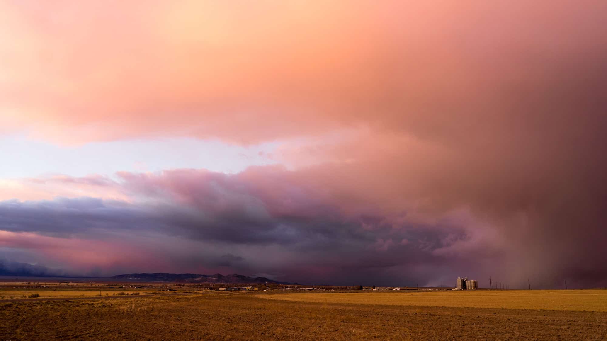 A dramatic sunset sky with sweeping pink and purple clouds looms over a flat, dry plain in Beaver County, Utah, with a distant grain elevator and faint outlines of mountains on the horizon.