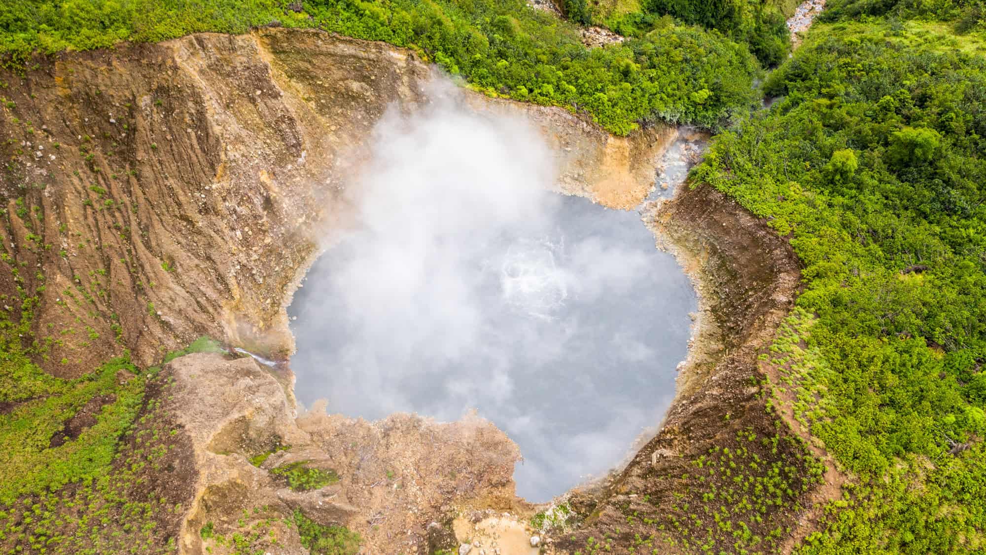 Aerial image of Dominica’s Boiling Lake shows a massive, steaming volcanic crater surrounded by lush green forest and rocky terrain, with thick plumes of vapor rising from the bubbling grey water.