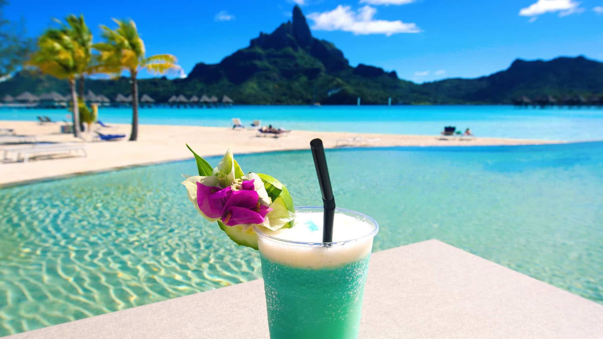 a tropical blue drink sits in the foreground with the landscape of bora bora in the background along with the incredible blue ocean