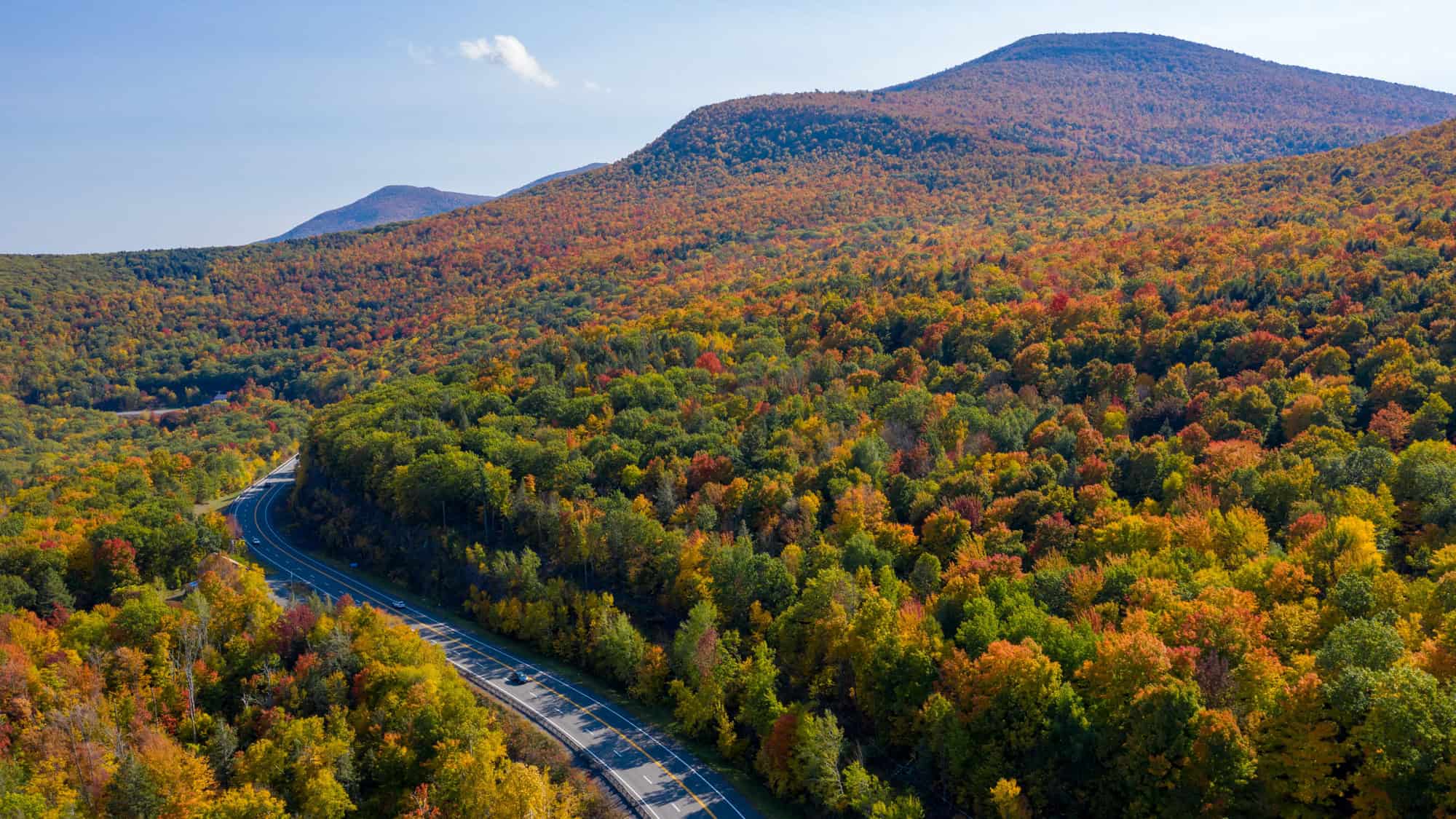 A winding highway cuts through the vibrant fall foliage of the Catskill Mountains in New York, with red, orange, and yellow trees carpeting the rolling hills.