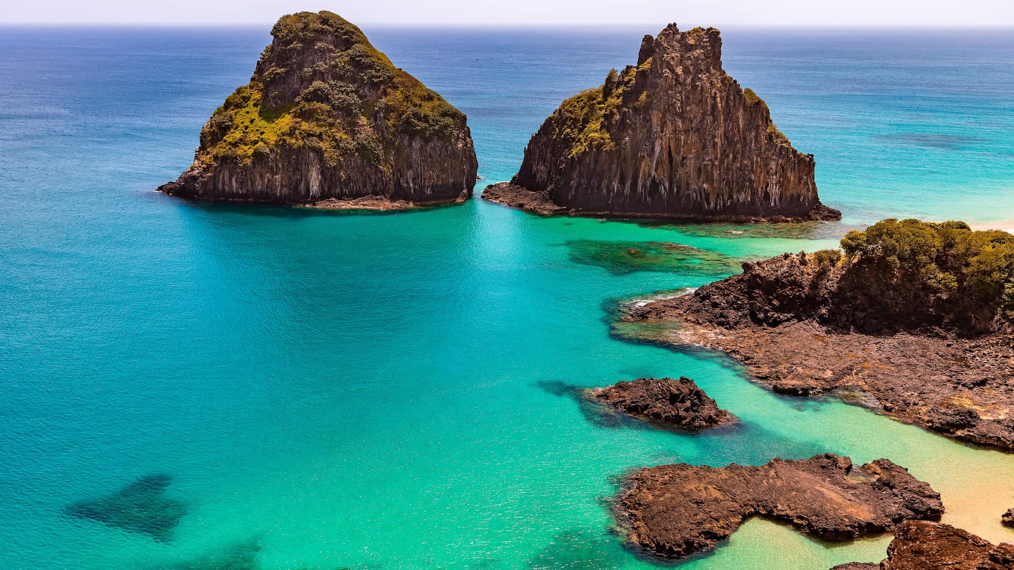 Aerial view of two rocky islands surrounded by clear turquoise waters and green foliage, part of the archipelago of Fernando de Noronha, Brazil.