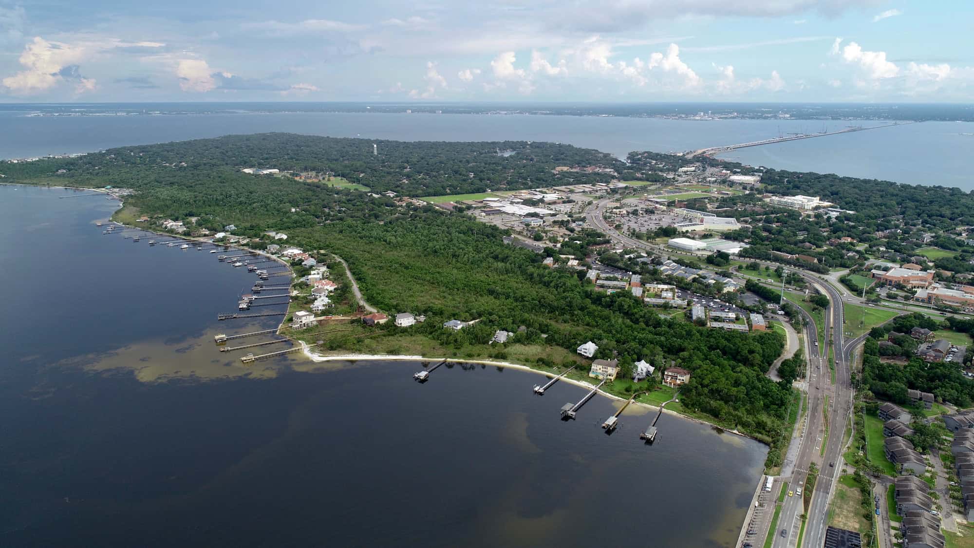 Aerial shot of Gulf Breeze, Florida, showing a mix of residential docks and urban development bordered by dense green trees and calm bay waters stretching toward the Pensacola Beach bridge.