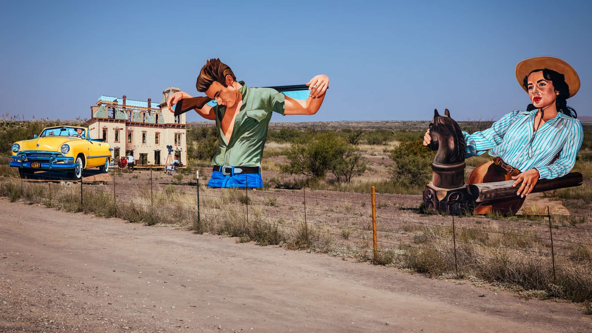 Outdoor art installation in Marfa, Texas, featuring large cutouts of James Dean and Elizabeth Taylor alongside a vintage yellow convertible and a replica of a Western film set building from the movie Giant.