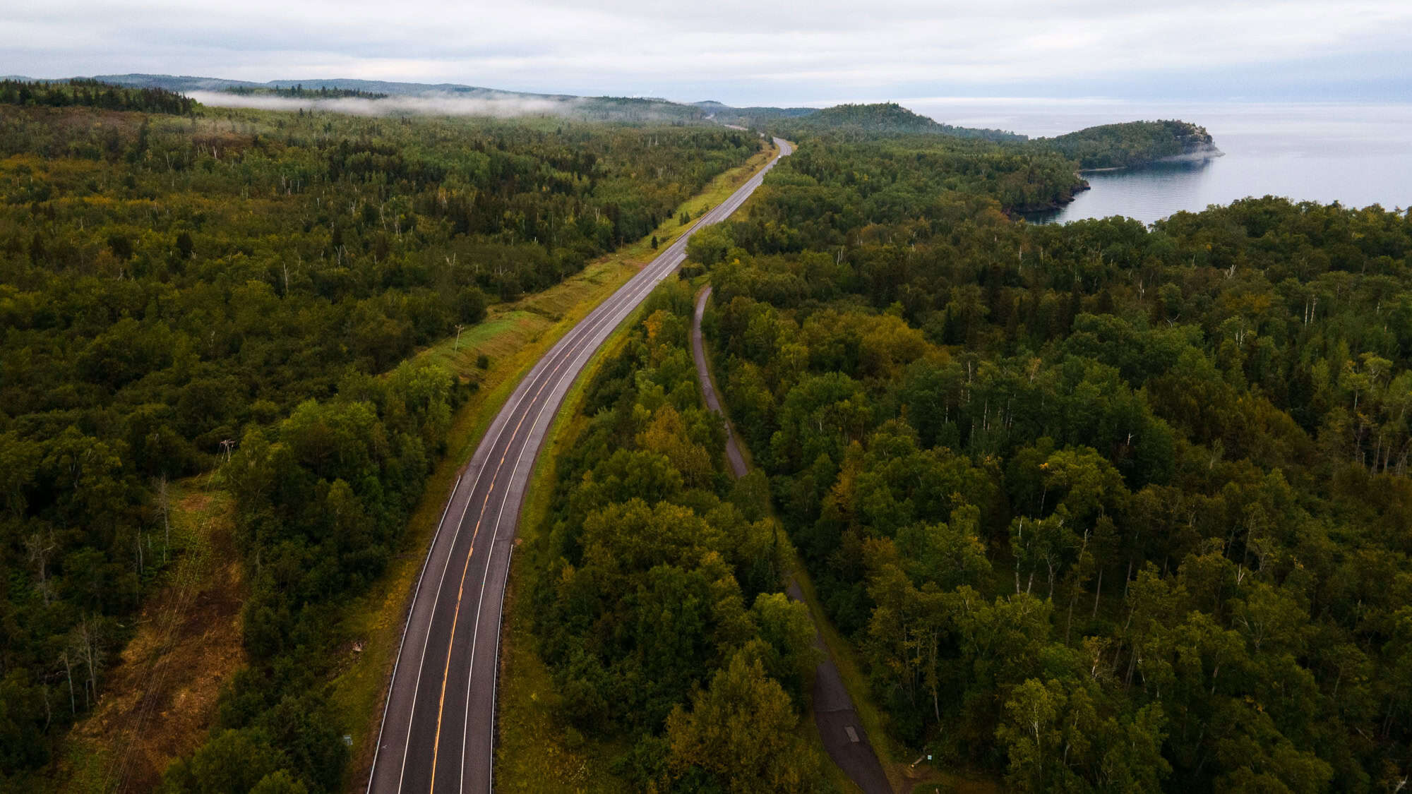 An aerial view of the North Shore Scenic Drive shows the highway slicing through a lush green forest beside Lake Superior, with fog rolling gently over distant hills and a rugged peninsula jutting into the lake.