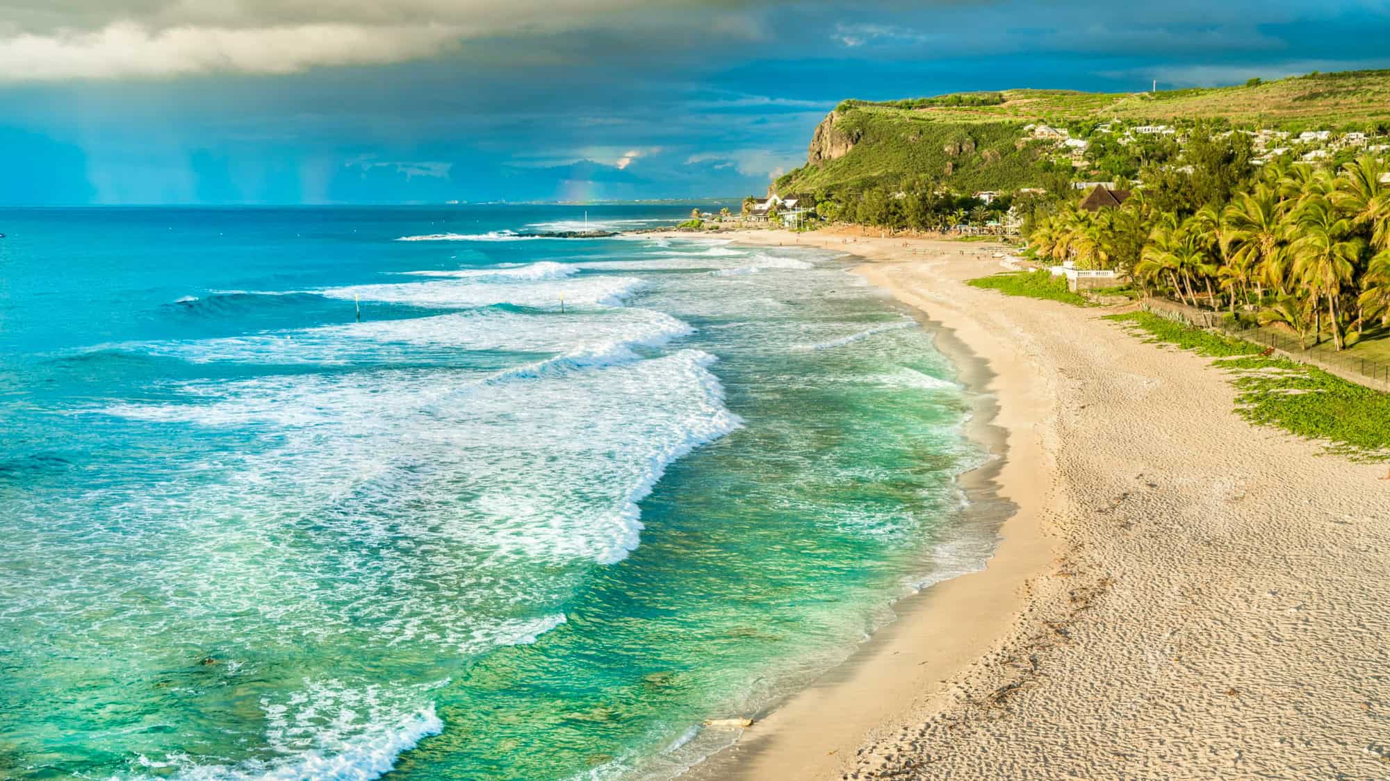 Scenic coastline of Reunion Island with waves crashing against the sandy shore and storm clouds in the distance.