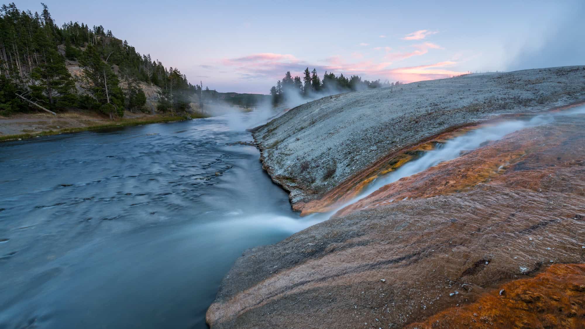 A steaming river with mineral deposits on its banks in Yellowstone National Park, with a serene pink and blue sky at dawn.