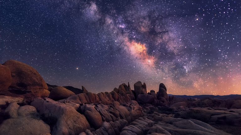 A starry night over the rocky desert landscape of Joshua Tree National Park, with the Milky Way galaxy vividly visible in the sky.