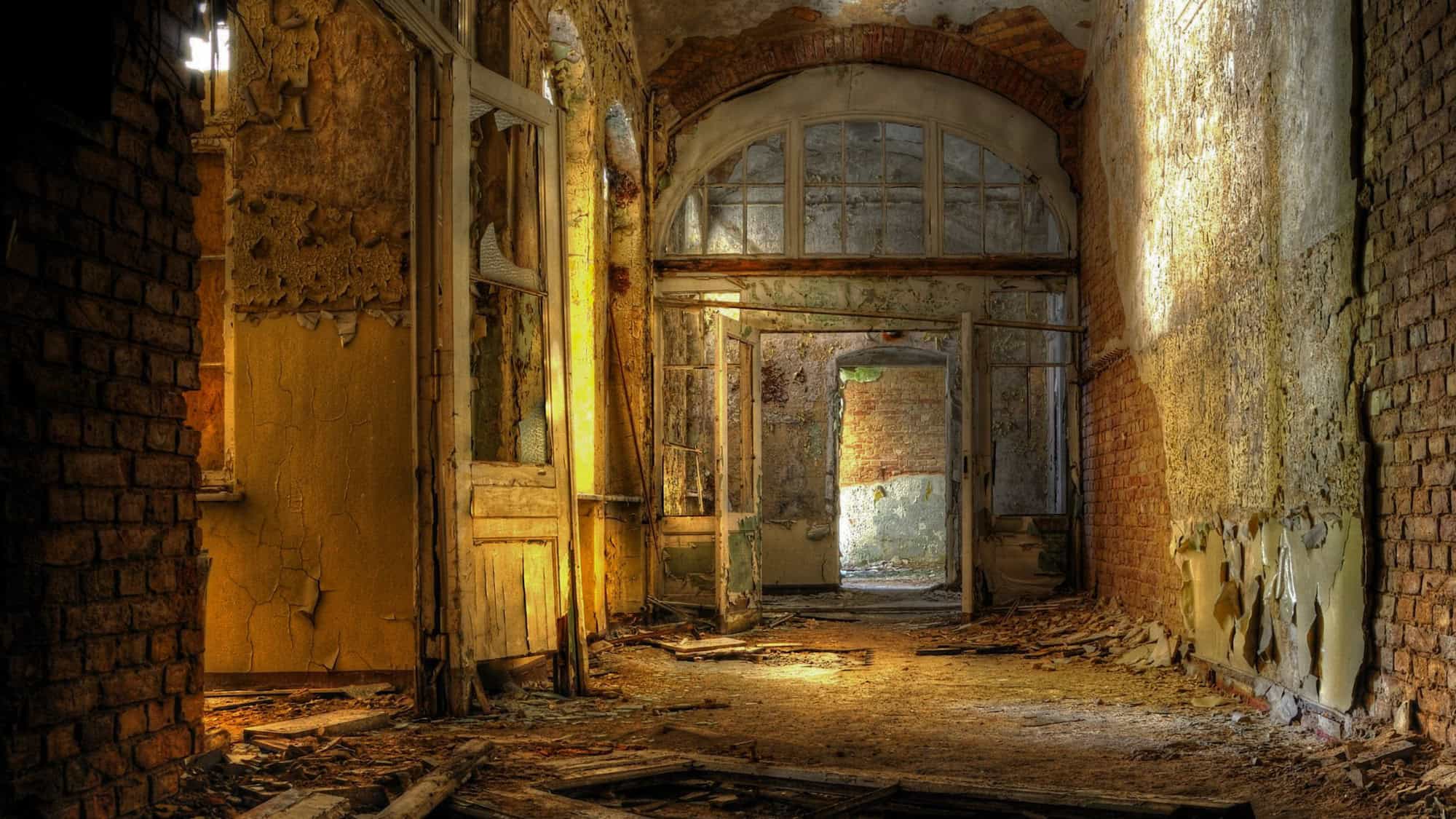 An abandoned hallway with peeling paint, crumbling walls, and broken doors in the Beelitz-Heilstätten hospital. The shafts of light filtering through the windows cast a haunting glow on the deteriorating structure.