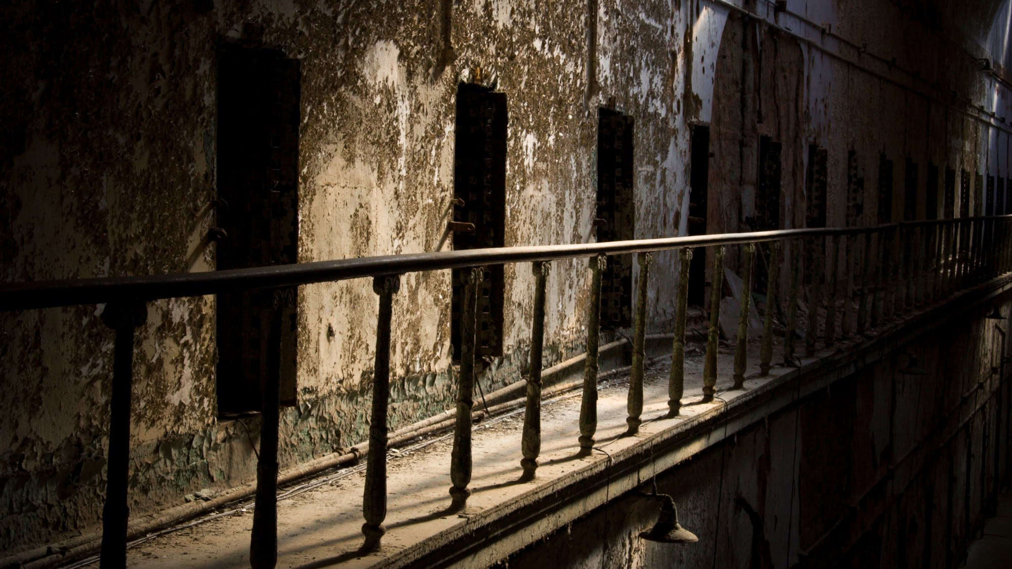 An eerie, dimly lit prison corridor with peeling paint and decaying walls. The dark atmosphere highlights the abandoned and deteriorating state of the facility.