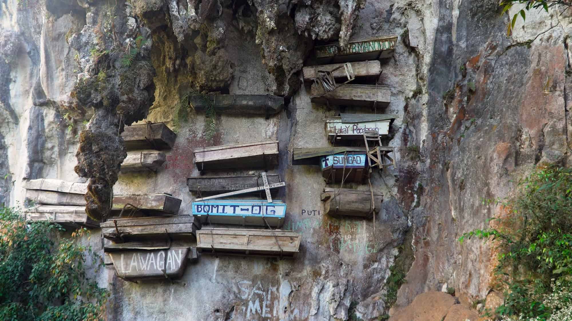 Several wooden coffins suspended on a cliffside, a tradition of the indigenous people. The coffins are weathered and precariously placed, creating a dramatic and eerie visual against the rock face.