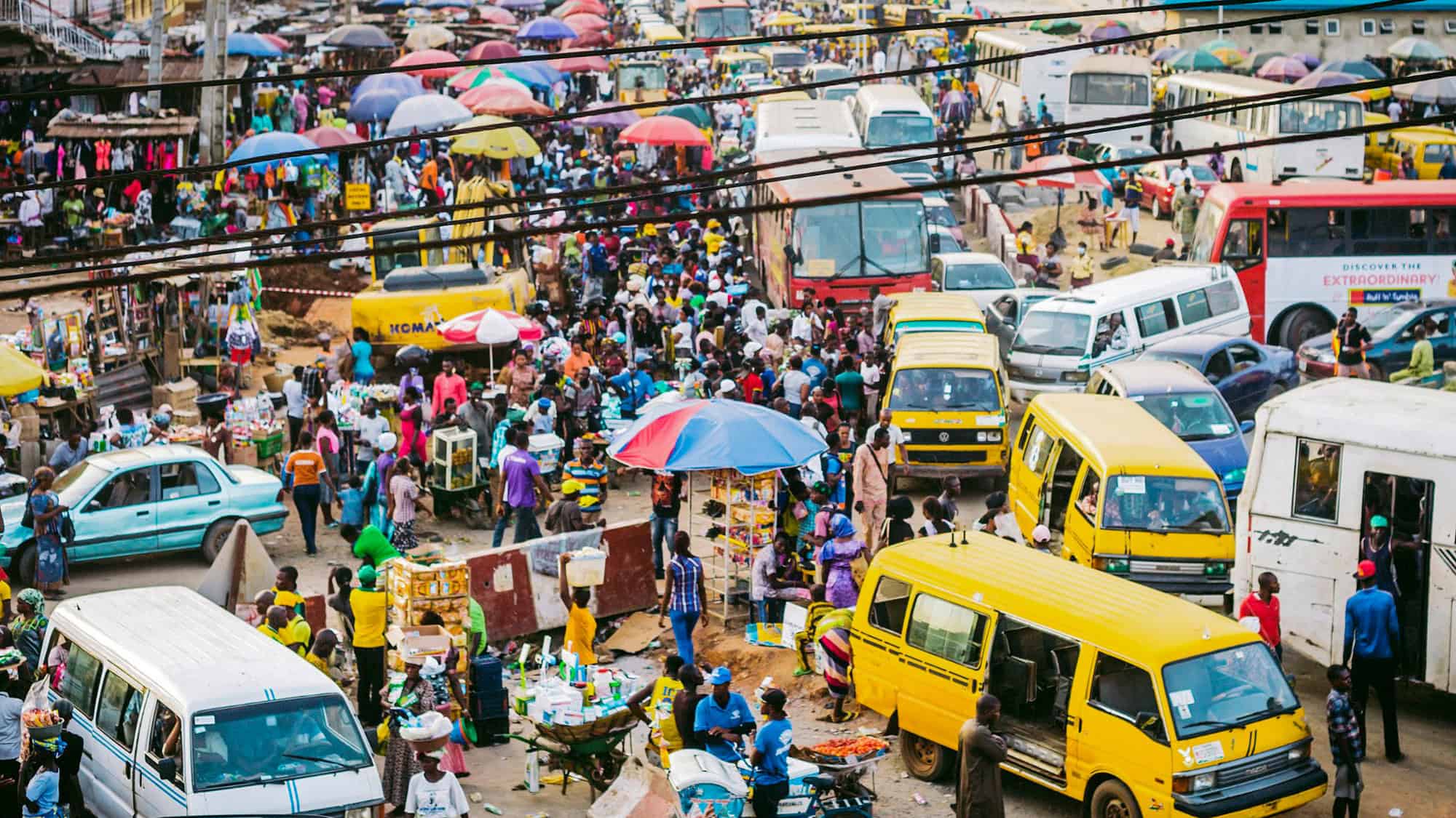 A crowded marketplace with yellow buses, cars, and pedestrians under colorful umbrellas. The scene is vibrant and bustling, reflecting the city's lively street market culture.