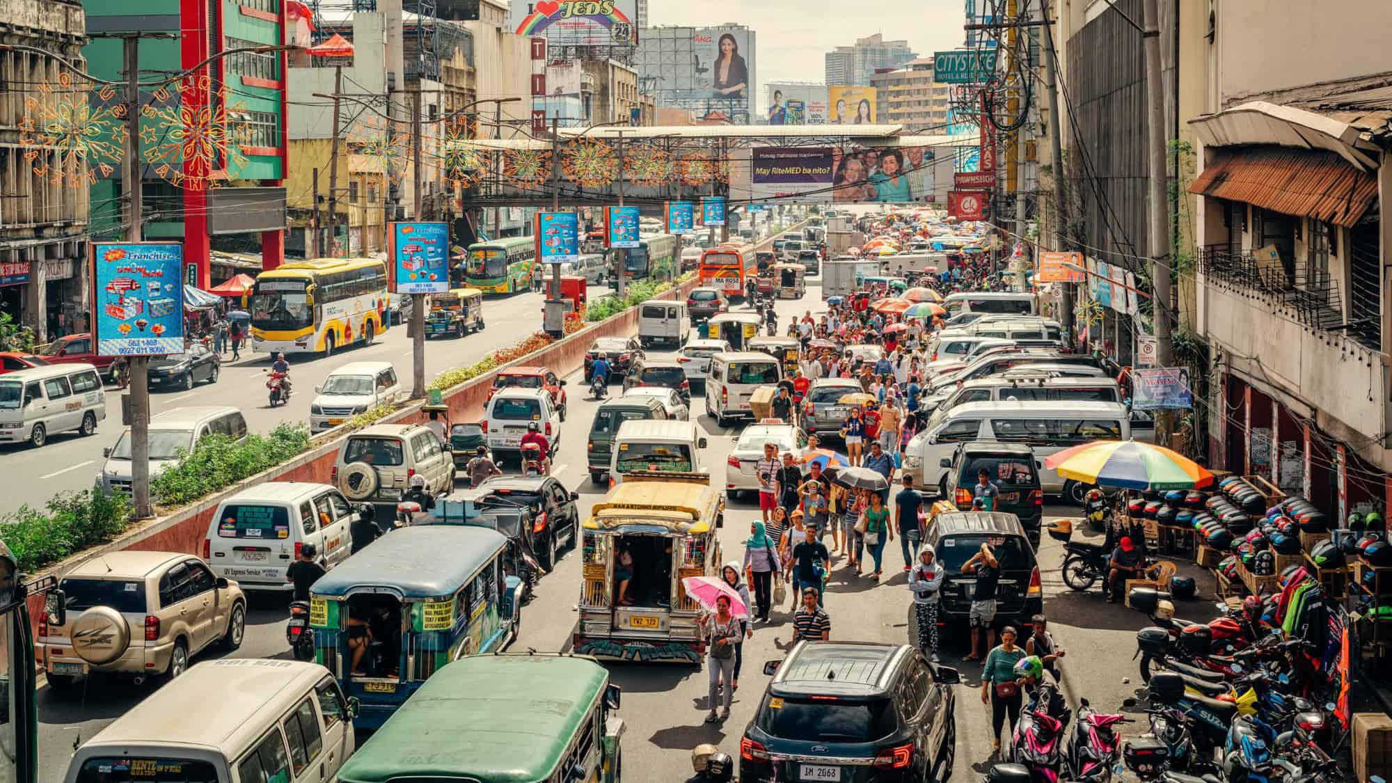 A busy street in Manila with vehicles like jeepneys and buses, surrounded by market vendors and colorful advertisements in the background.