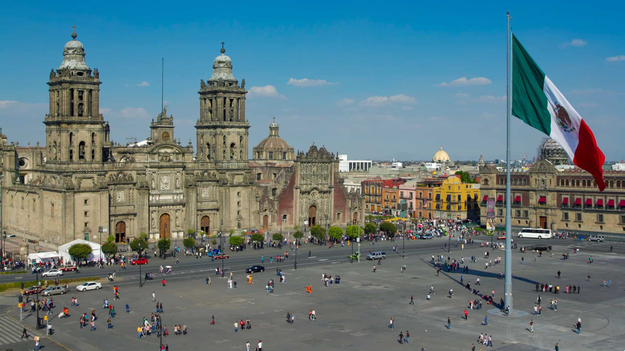 A bustling plaza in front of the Mexico City Metropolitan Cathedral, with a large Mexican flag waving prominently on the right.