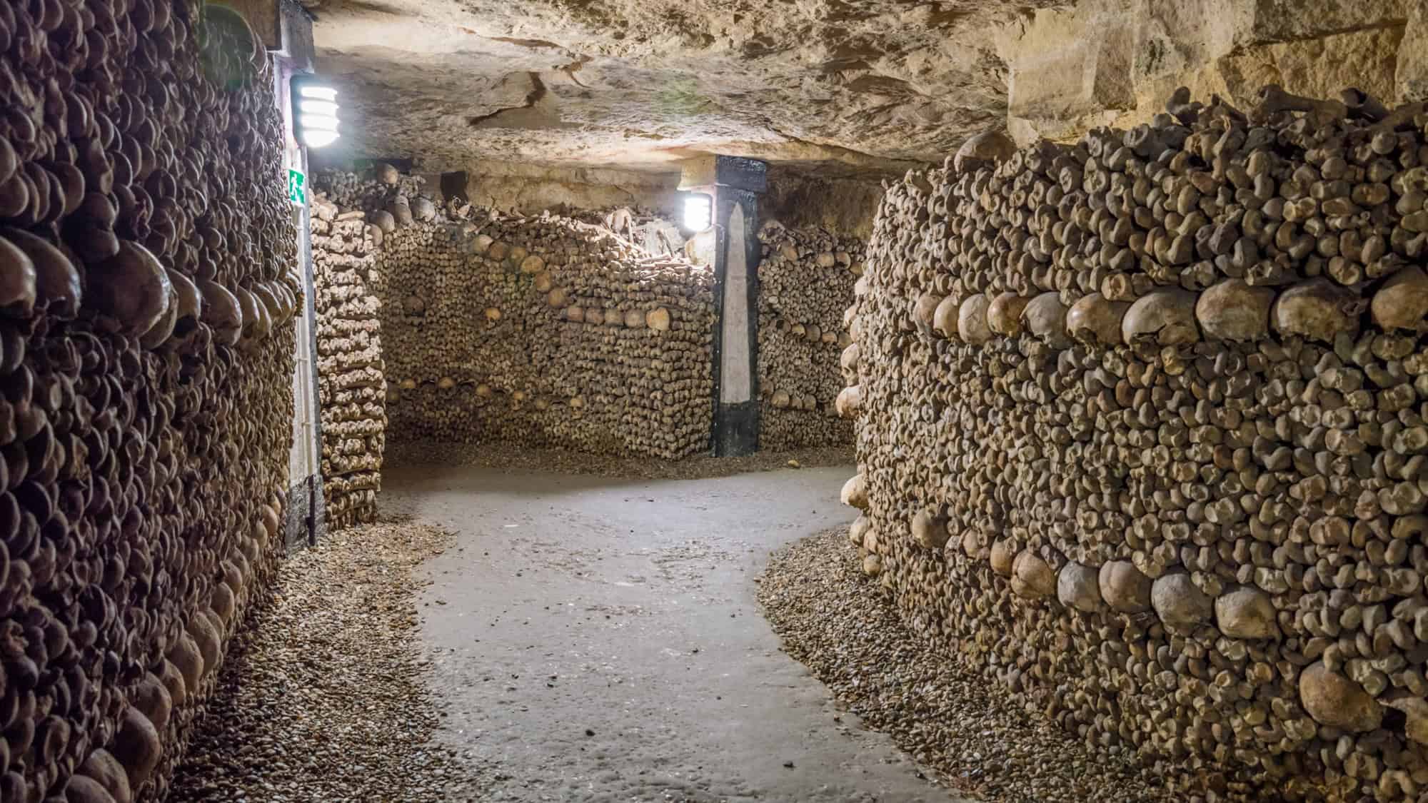 A narrow underground tunnel lined with thousands of human skulls and bones, stacked neatly along the walls of the Paris Catacombs, creating a haunting, yet organized, labyrinth of the dead.