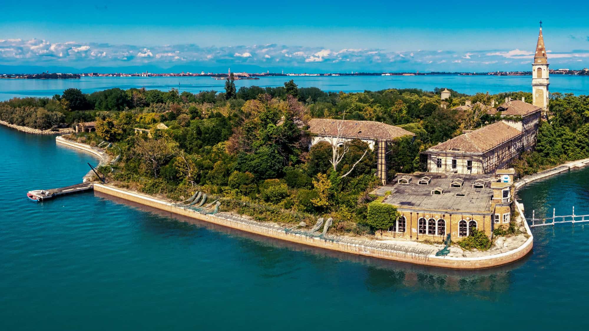 Aerial view of a seemingly peaceful but overgrown and abandoned island, Poveglia, with old, dilapidated buildings and lush greenery set against a bright blue sea.