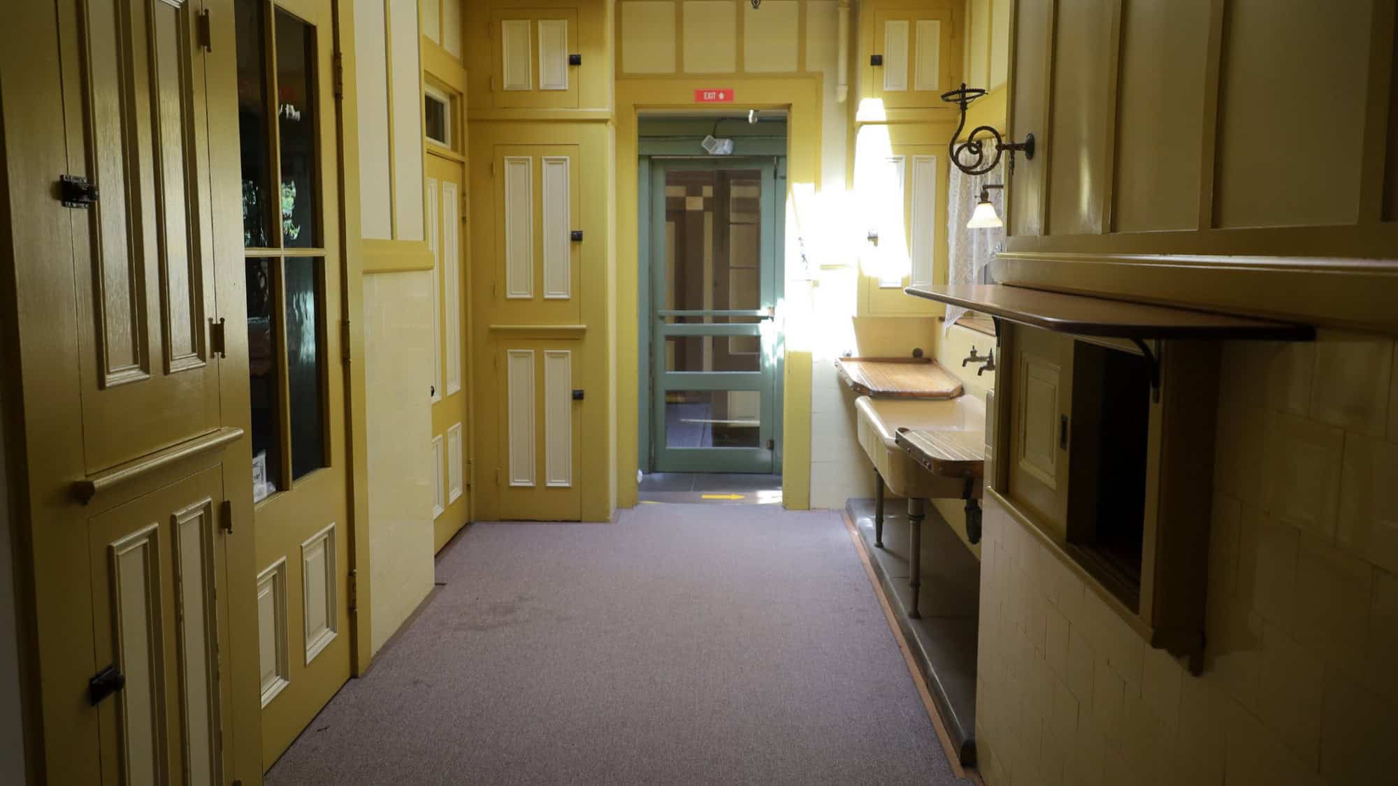 An old, yellow-painted hallway with tall, paneled doors and a vintage feel. The hallway has wooden countertops and light streaming in from the open door, creating a sense of isolation and mystery.