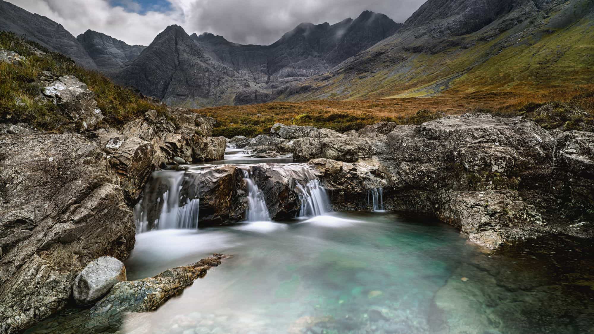 Several small waterfalls pouring into a pool with a beautiful rocky mountain backdrop in Scotland.