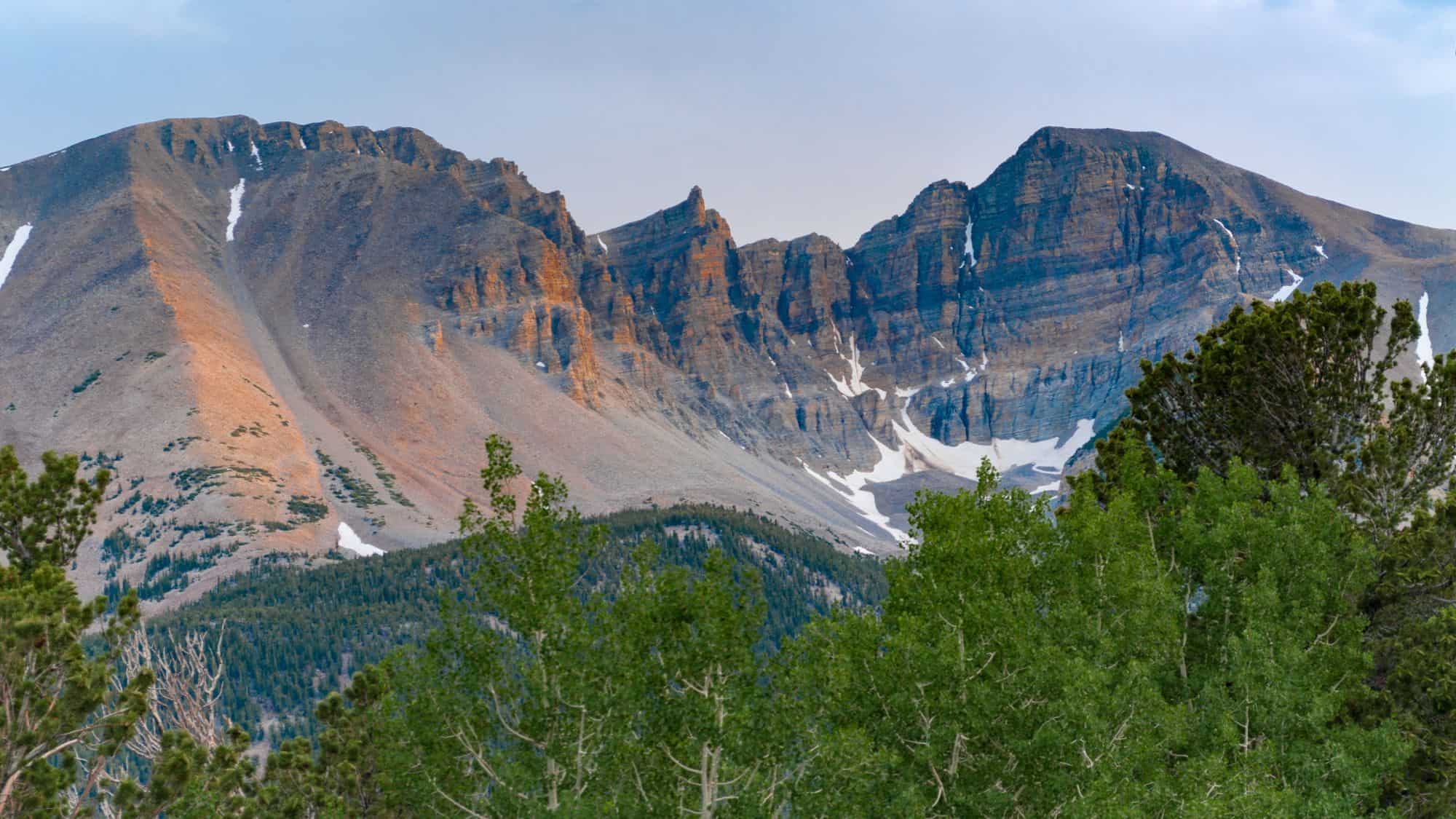 A view of Great Basin National Park with its rocky mountains standing tall with a bit of snow behind green trees.