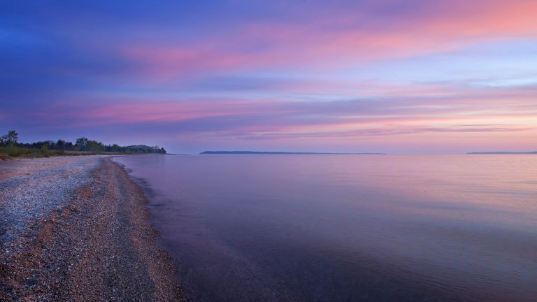 A serene shoreline scene during dawn, with a calm lake reflecting the soft hues of a purple and pink sunrise. The quiet pebbled beach stretches into the distance under a pastel sky.