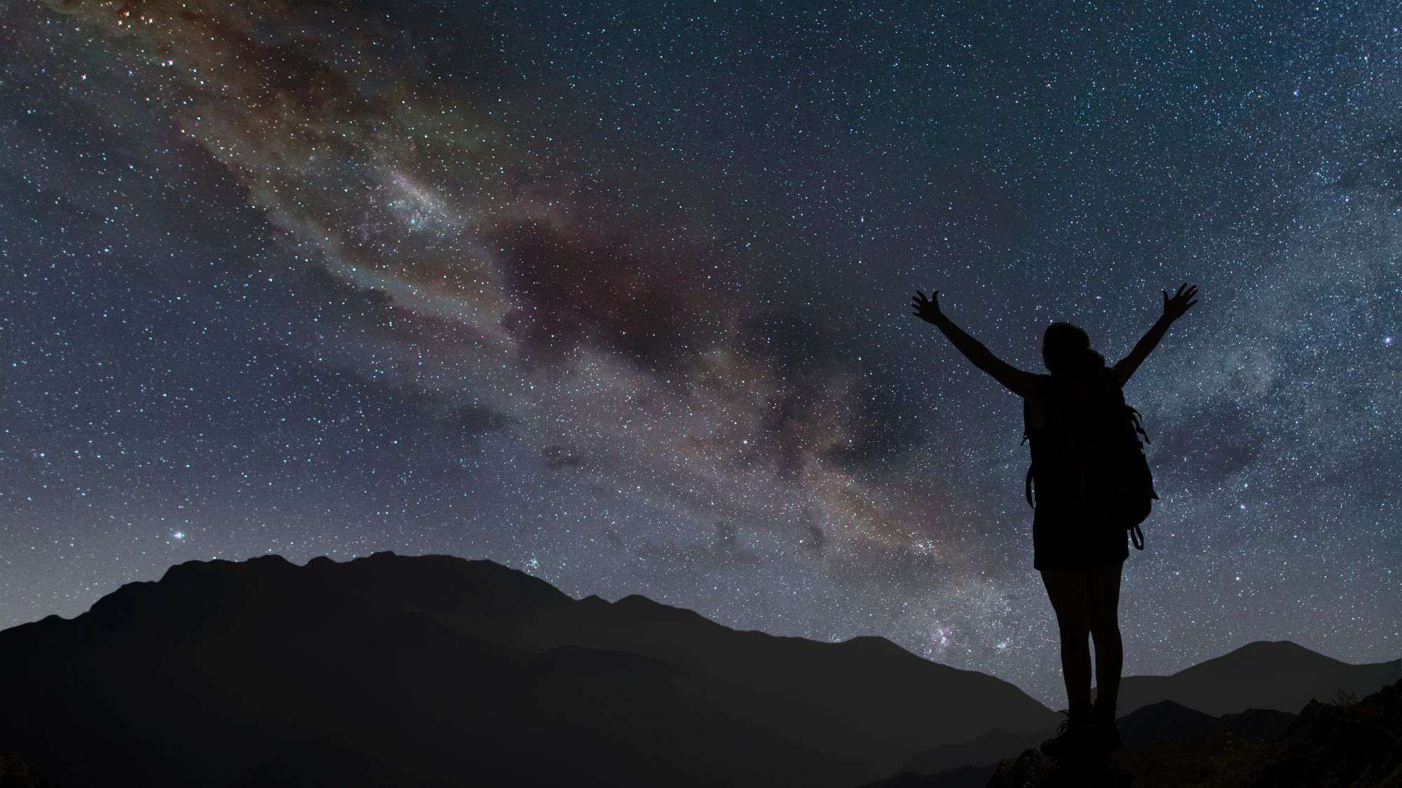 A silhouetted woman with her arms raised stands under the milky way in a mountain landscape.