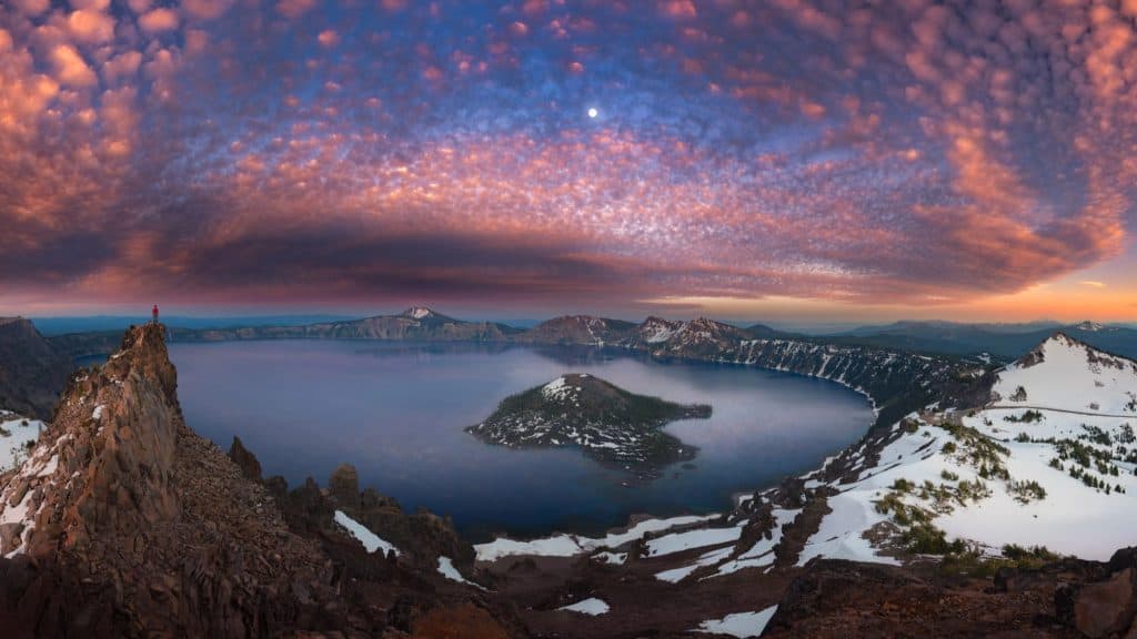 A panoramic view of Crater Lake at sunrise or sunset, with vibrant clouds reflecting over the calm water and snowy patches scattered on the rocky terrain.