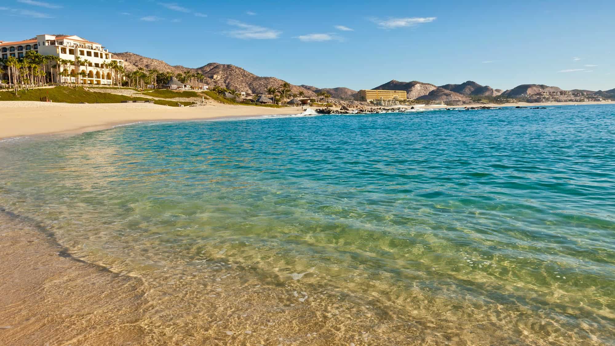A calm beach at Cabo San Lucas, with clear, shallow turquoise water gently lapping against the sandy shore. Hills dotted with resorts are visible in the distance under a bright blue sky.