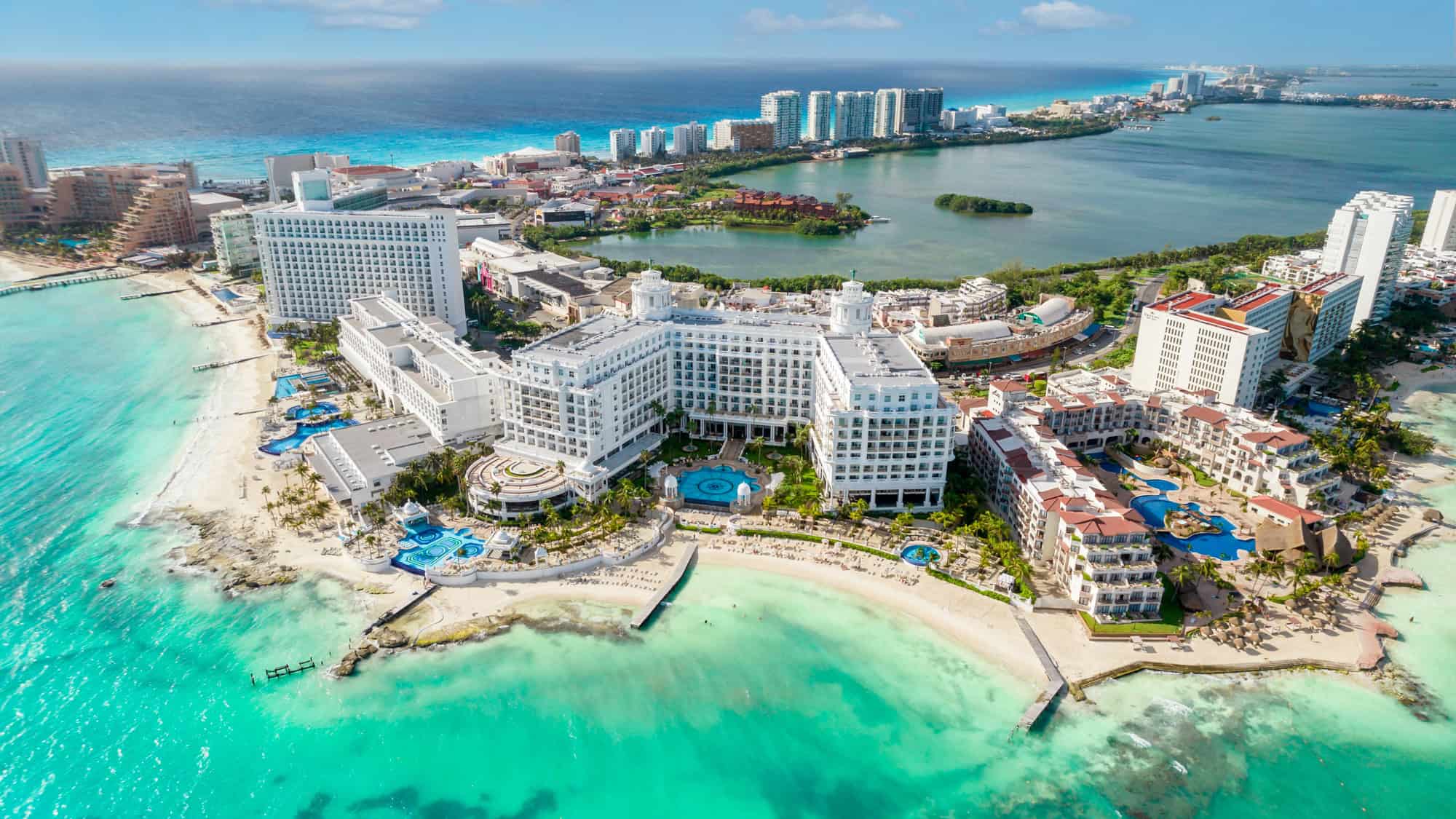 An aerial view of Cancun's Hotel Zone, showcasing luxury resorts lining a pristine white sandy beach with vibrant turquoise water. The cityscape and lagoon are visible in the background, emphasizing the blend of urban and natural beauty.