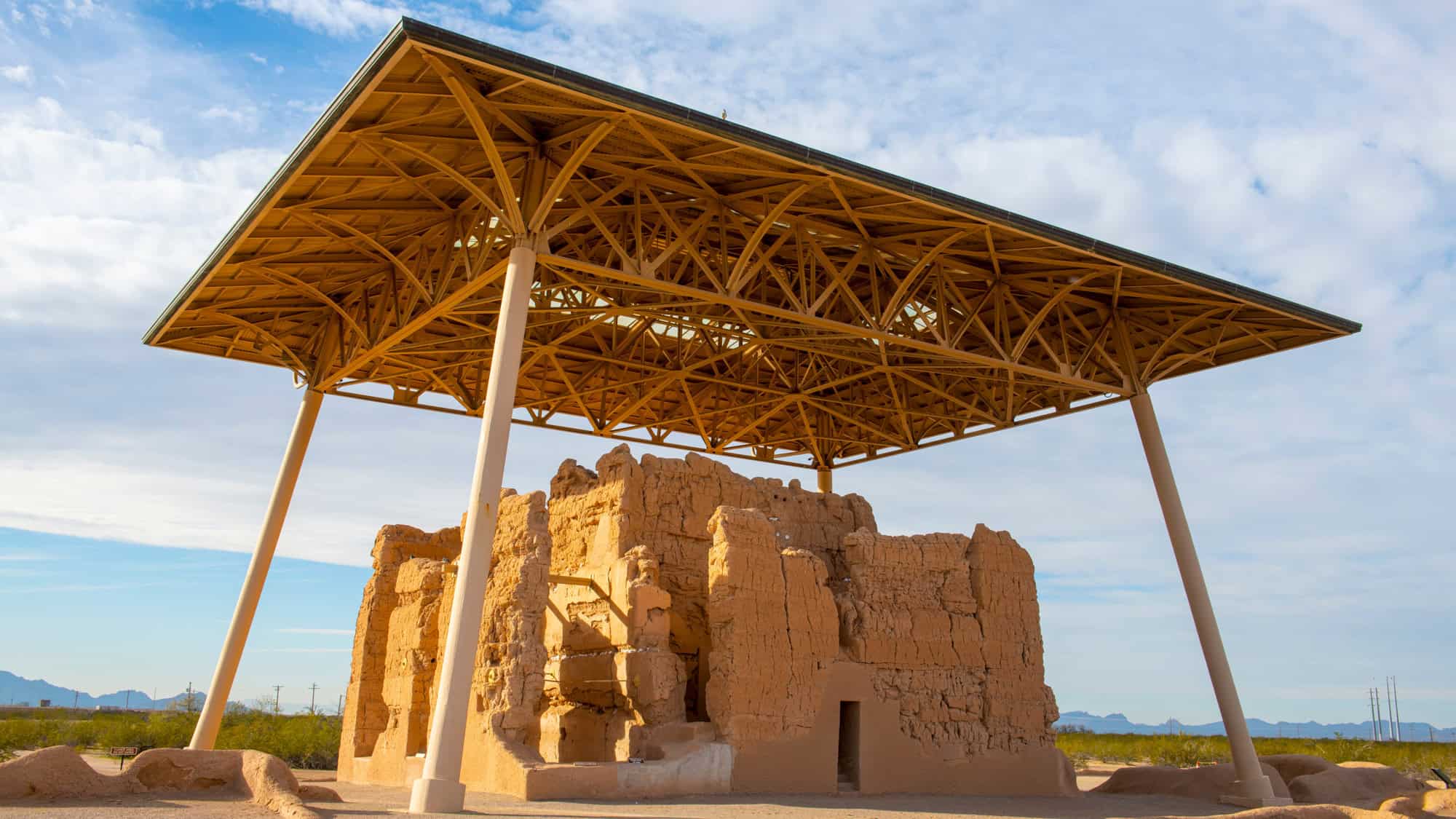 An ancient Hohokam adobe structure stands beneath a protective steel canopy, surrounded by desert landscape and distant mountains in southern Arizona.
