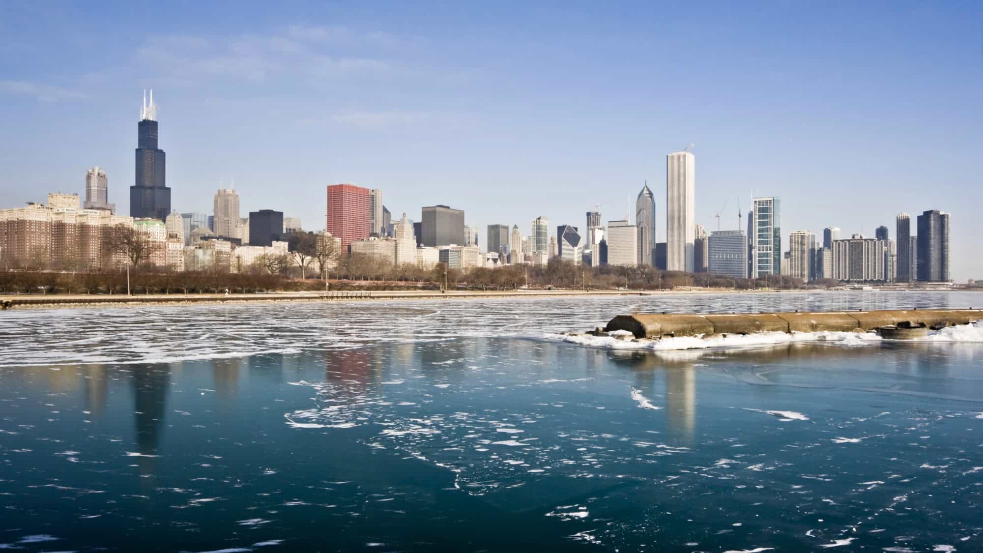 The Chicago skyline on a cold winter day, with icy Lake Michigan in the foreground and iconic skyscrapers like Willis Tower reflecting on the partially frozen water.