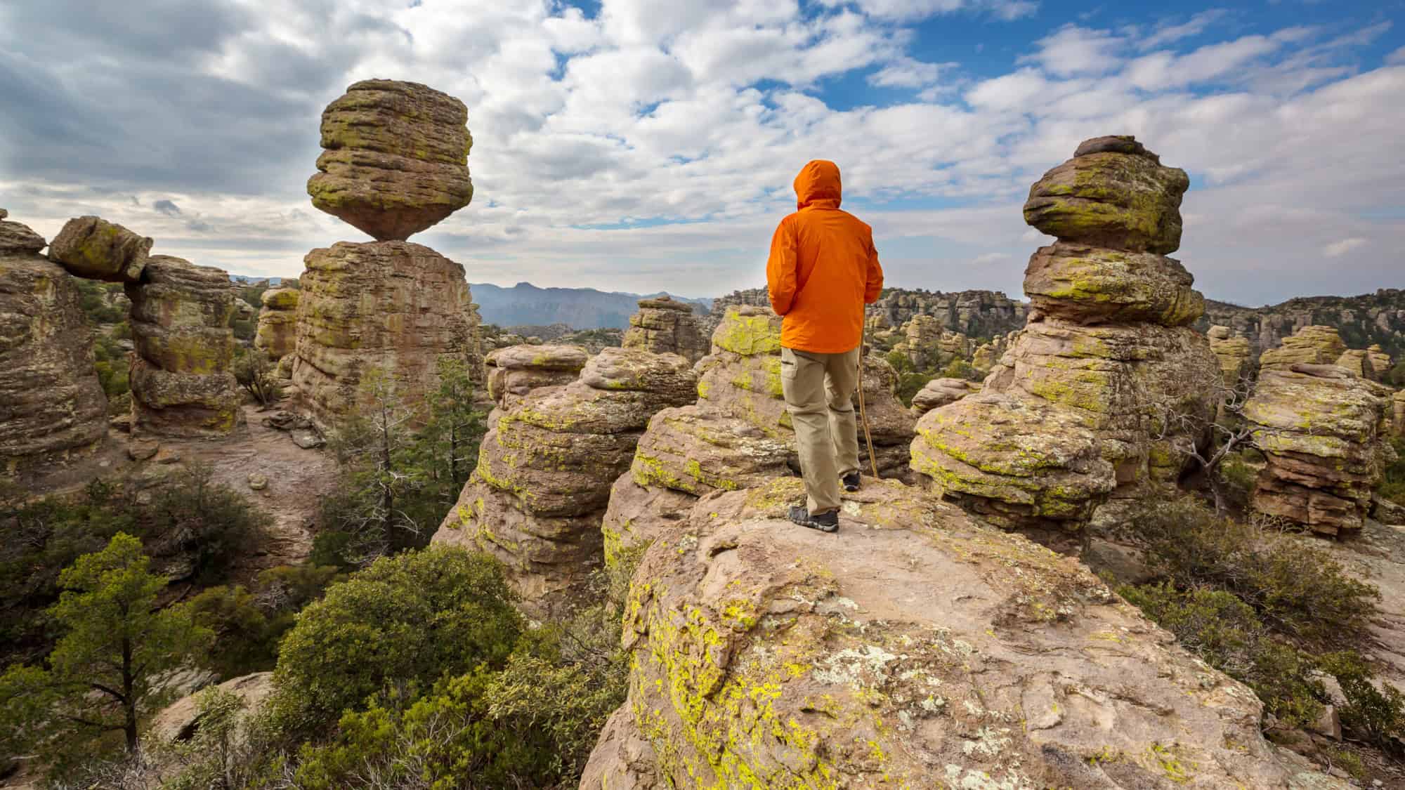 A hiker in an orange jacket overlooks a dramatic landscape of towering rock spires and massive balancing stones in Chiricahua National Monument, Arizona.