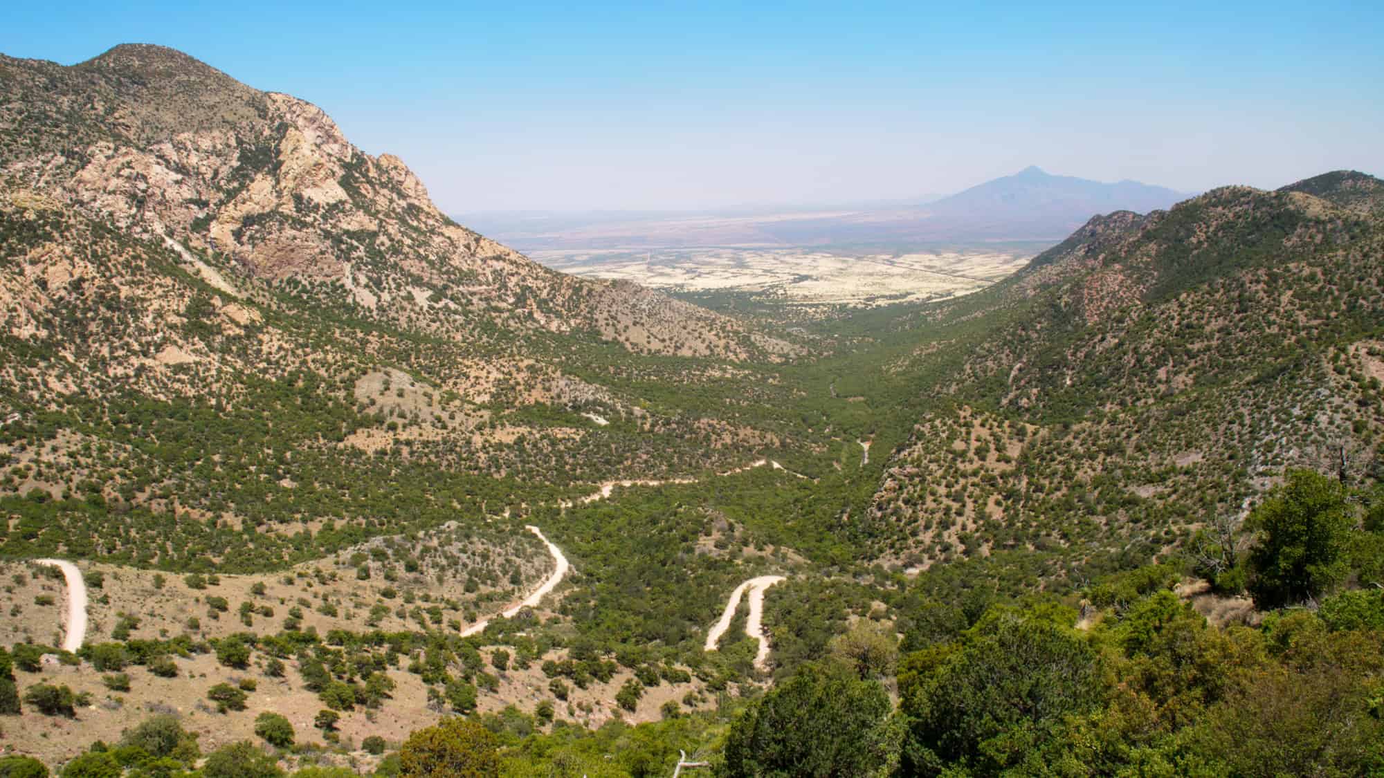 A winding dirt road snakes through a wide, green valley flanked by rugged desert mountains, offering a sweeping view toward the flatlands and distant peaks.