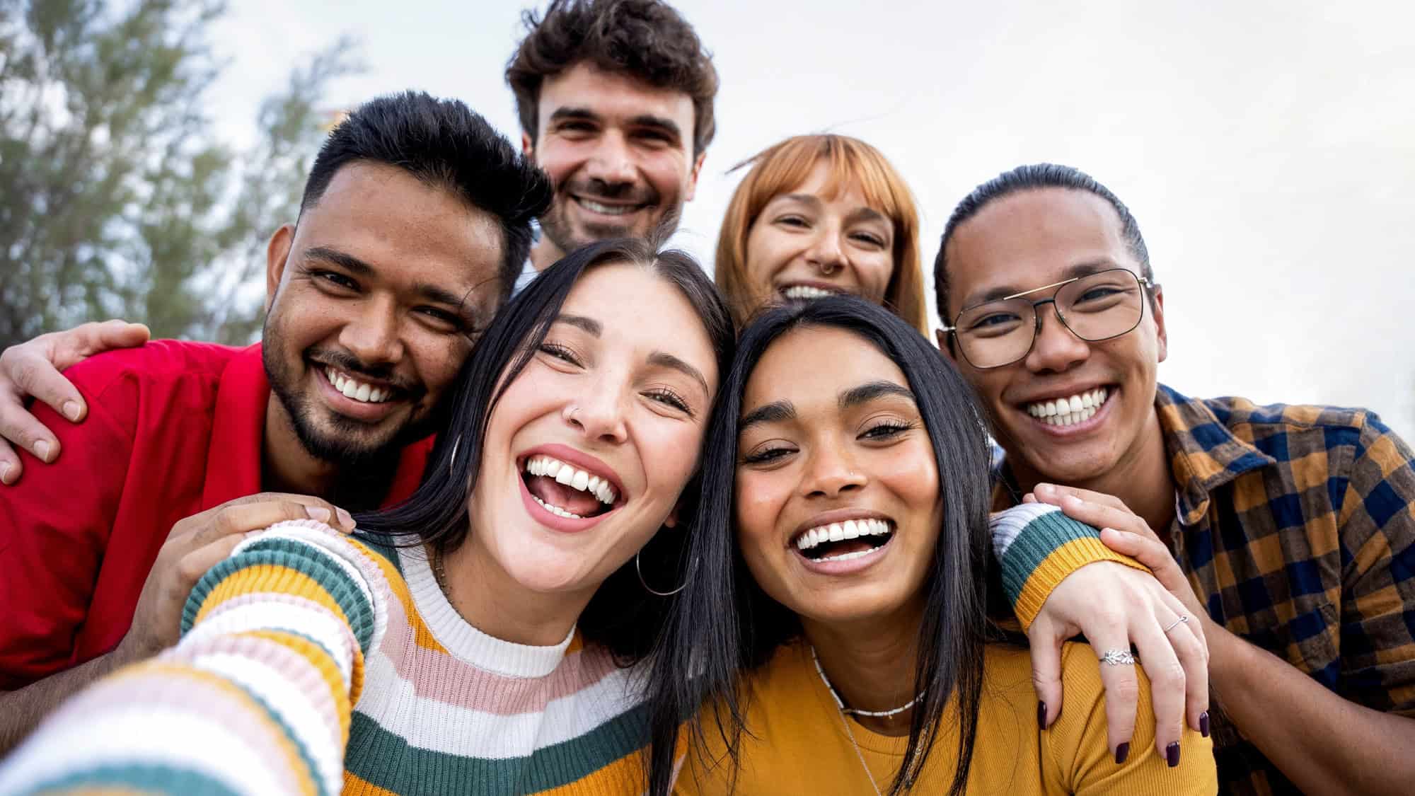A joyful group of six young adults poses closely for a selfie outdoors, all smiling brightly with arms around each other against a blurred natural background.
