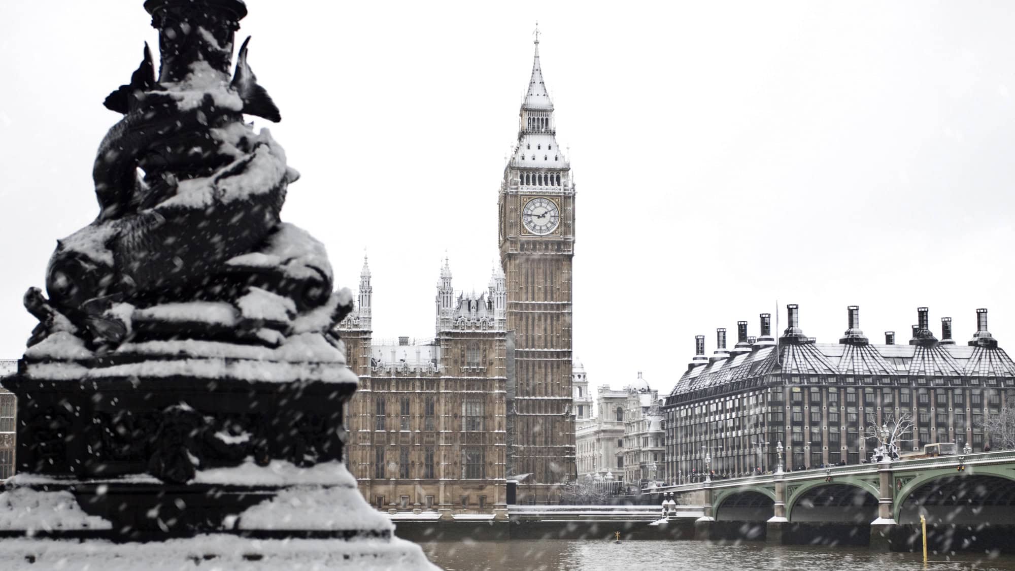 A snow-dusted view of Big Ben and the Houses of Parliament from across the Thames. Snowflakes are visible against the overcast sky, creating a peaceful winter scene.