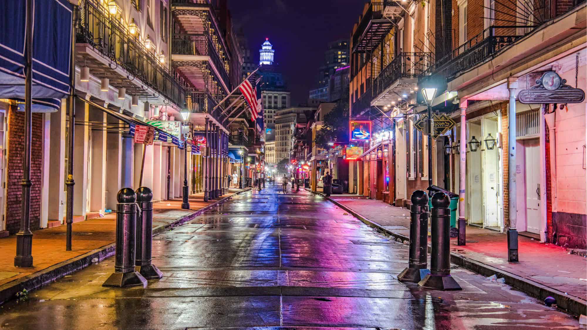 A vibrant night scene on New Orleans' Bourbon Street, with wet pavement reflecting the colorful neon lights of bars and restaurants lining the street, an American flag hanging above, and the faint glow of distant buildings.