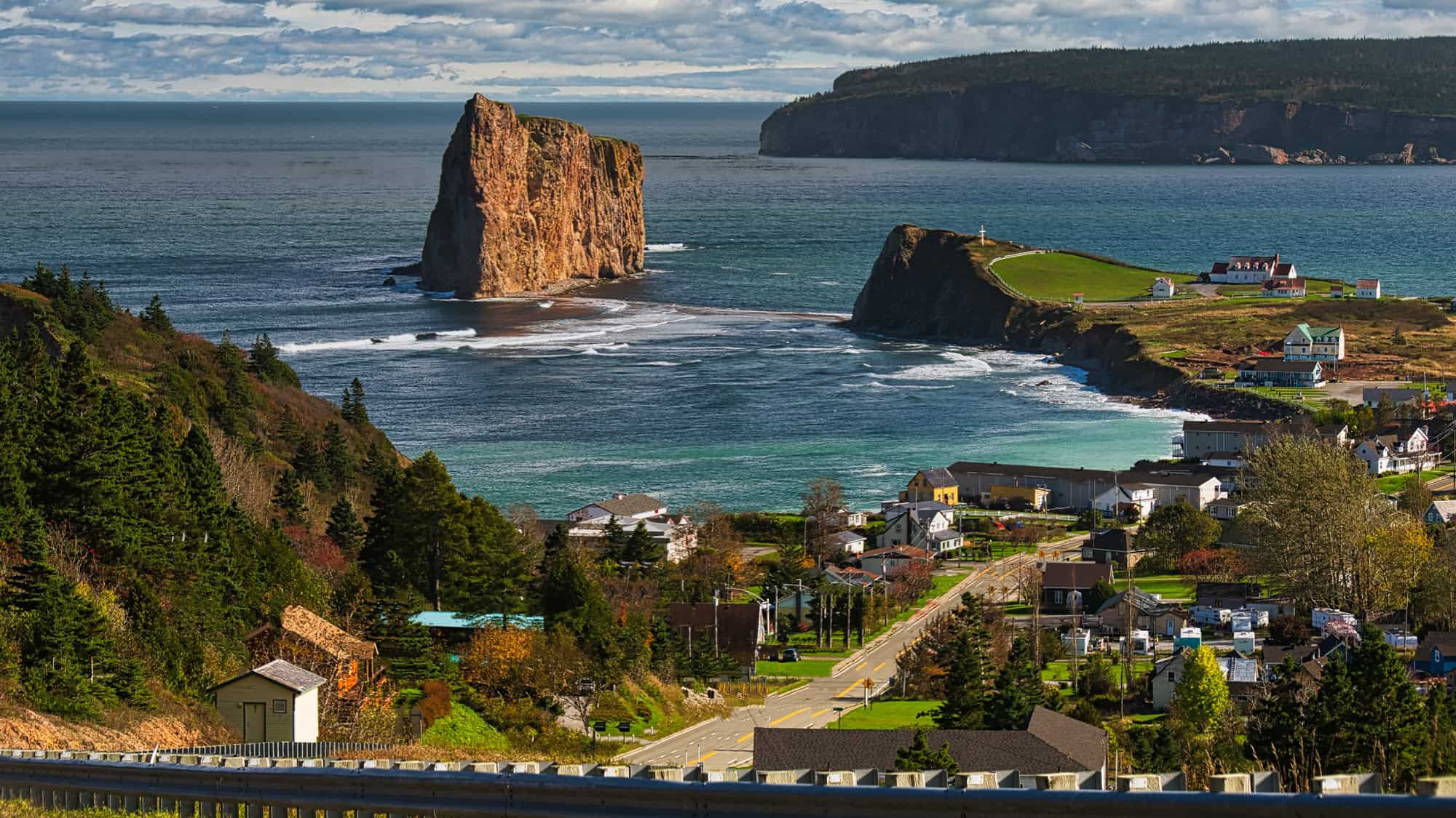 A dramatic coastal scene featuring Percé Rock, a massive limestone formation rising from the Gulf of Saint Lawrence near the shoreline of Percé, Quebec. The foreground shows a charming village with colorful houses, pine trees, and a winding road leading toward the cape and lighthouse.