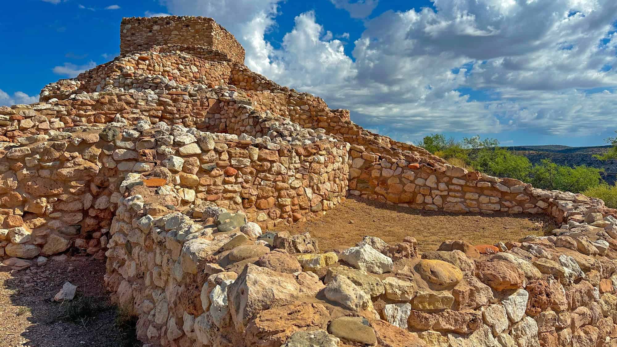 A terraced, hilltop stone ruin built by the Sinagua people features curved walls and stacked rock construction beneath a bright blue sky filled with puffy clouds.