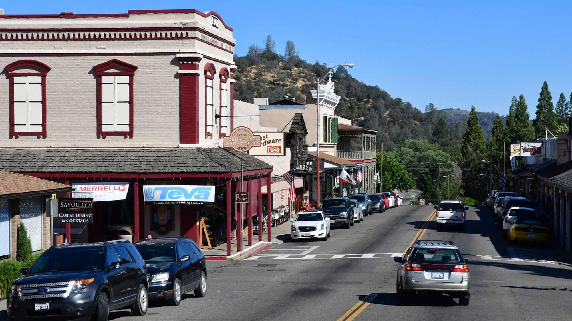 A charming small-town street with historic buildings and red-trimmed architecture, lined with parked cars and surrounded by hilly landscapes.