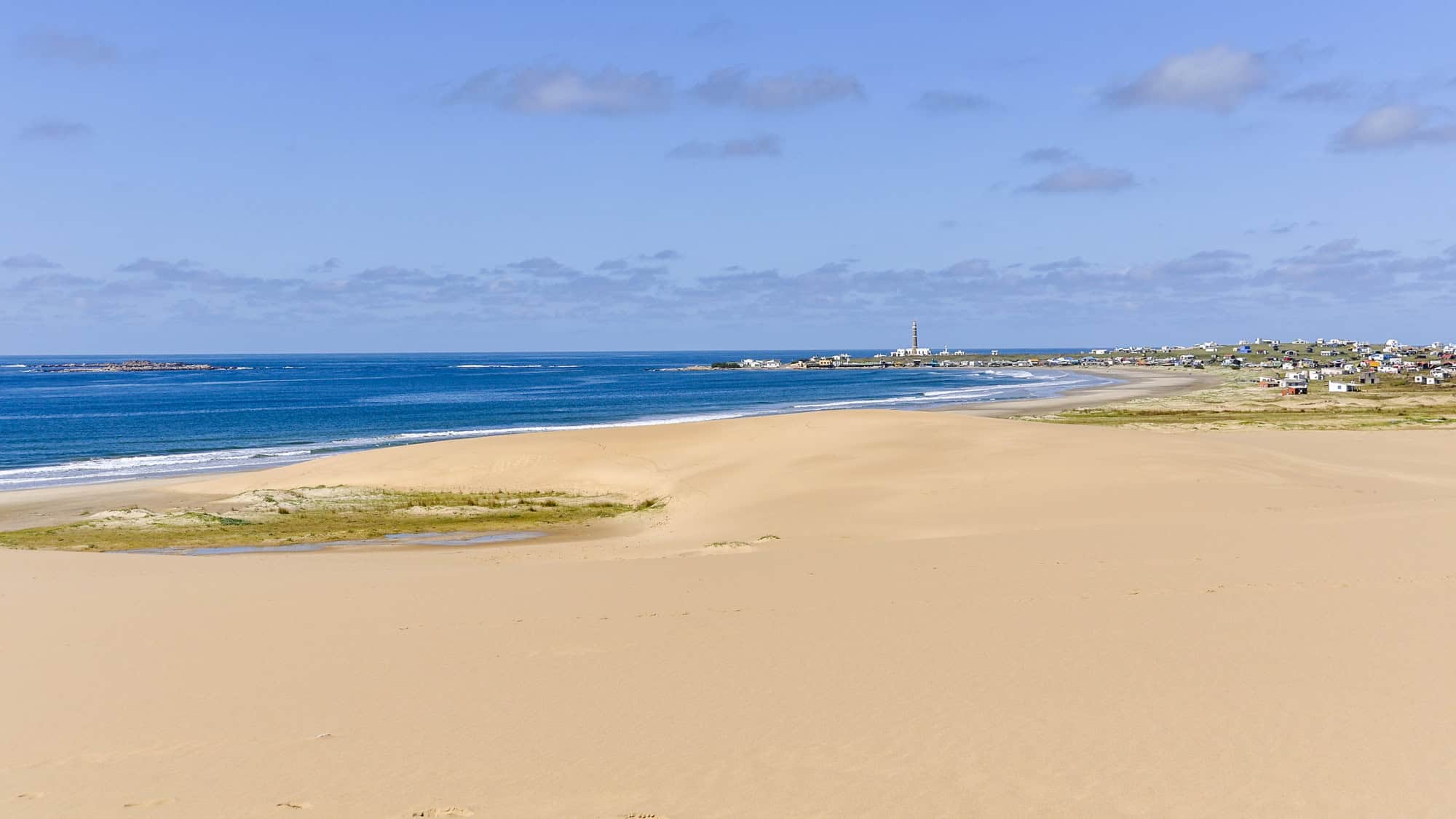 A wide stretch of golden sand dunes meets the deep blue Atlantic Ocean, with a distant village and lighthouse dotting the coastline under a clear sky.